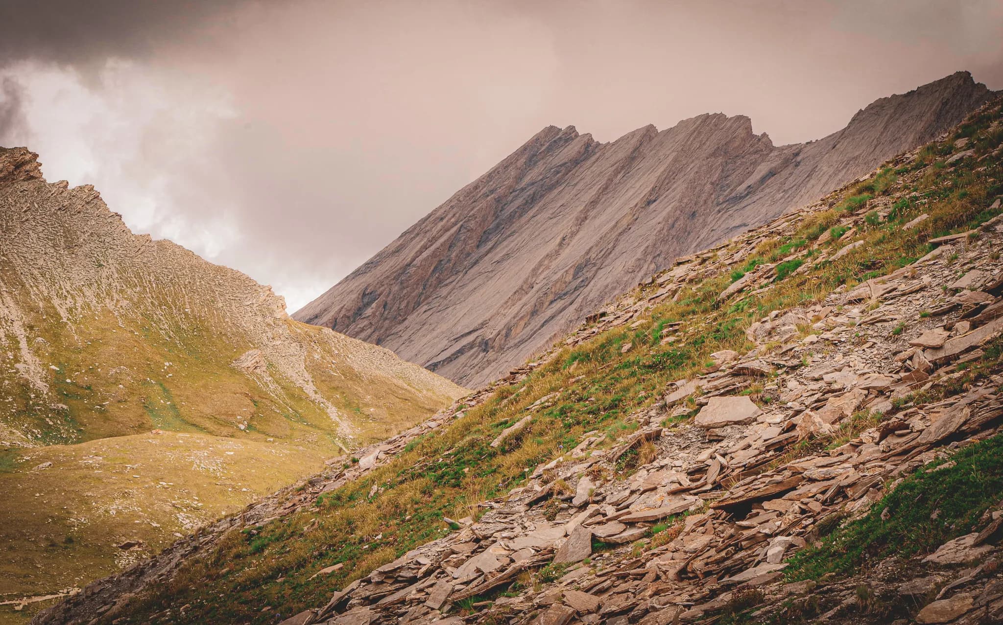 A mountainous landscape characterised by steep slopes and clear rock formations. The ground is strewn with flat stones and grasses, while rocky peaks tower above the scene, rising up to the horizon.