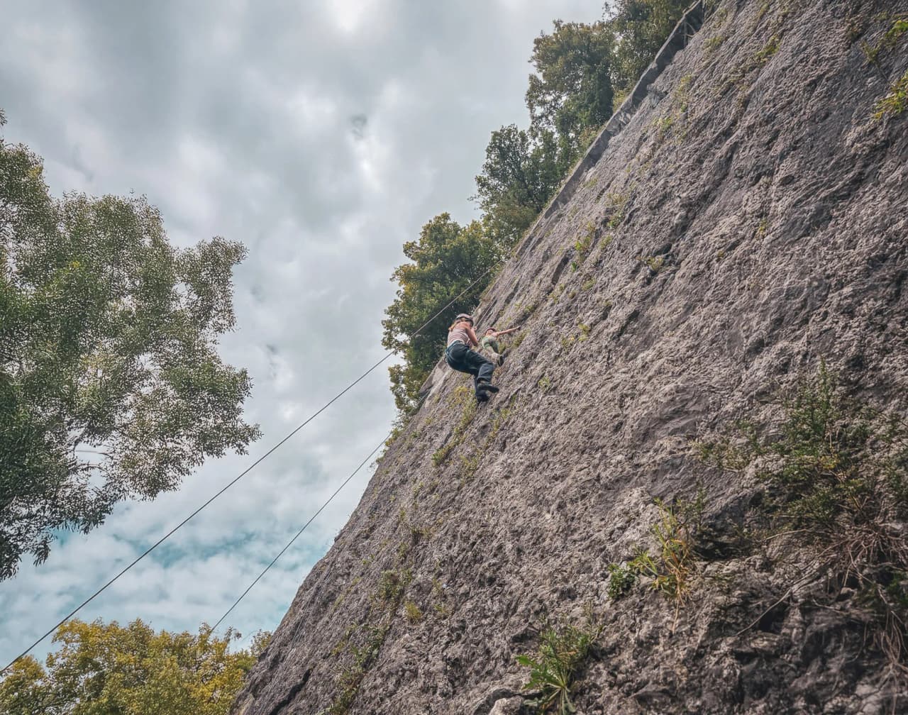 A climber ascends a cliff in Belgium, surrounded by greenery and cloudy skies.