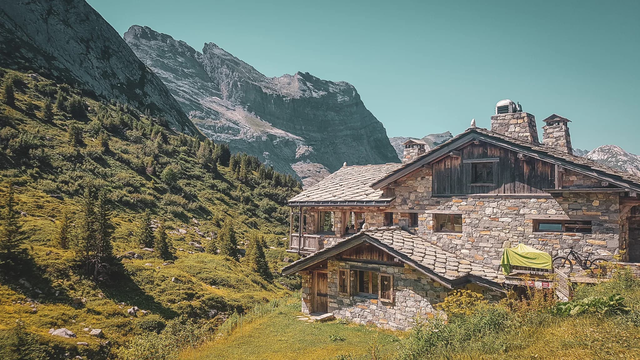 Un chalet en pierre élégant est niché au pied de majestueuses montagnes. Le bâtiment présente un toit en ardoise et des poutres en bois, créant un charme rustique. L'environnement est verdoyant avec des arbres et des plantes