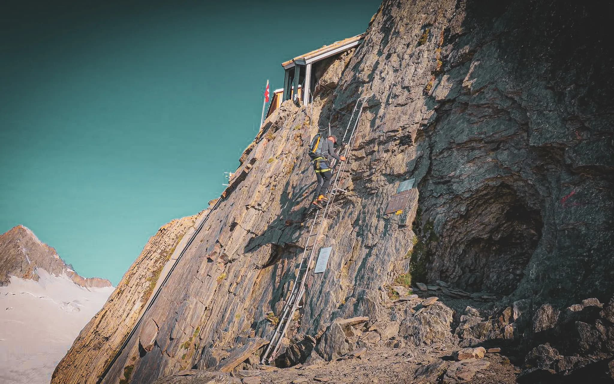 A climber climbs a steep rock face using a metal ladder, with a high-rise building visible above him. The landscape is mountainous, with beautiful light and clear skies overhead.