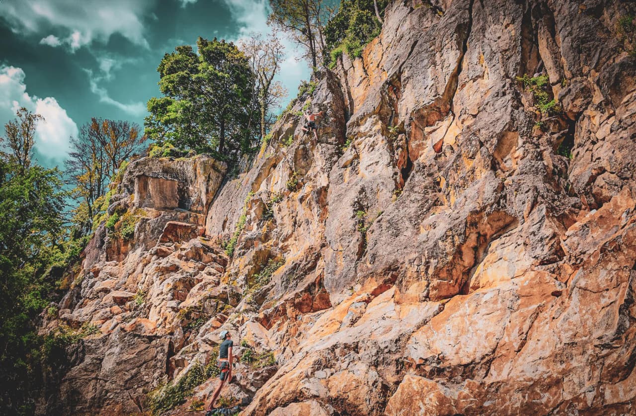 Un grimpeur est en train d'escalader une paroi rocheuse, entourée d'une végétation verdoyante. La falaise présente des roches de différentes textures et couleurs, allant du gris au beige. Au sommet de la