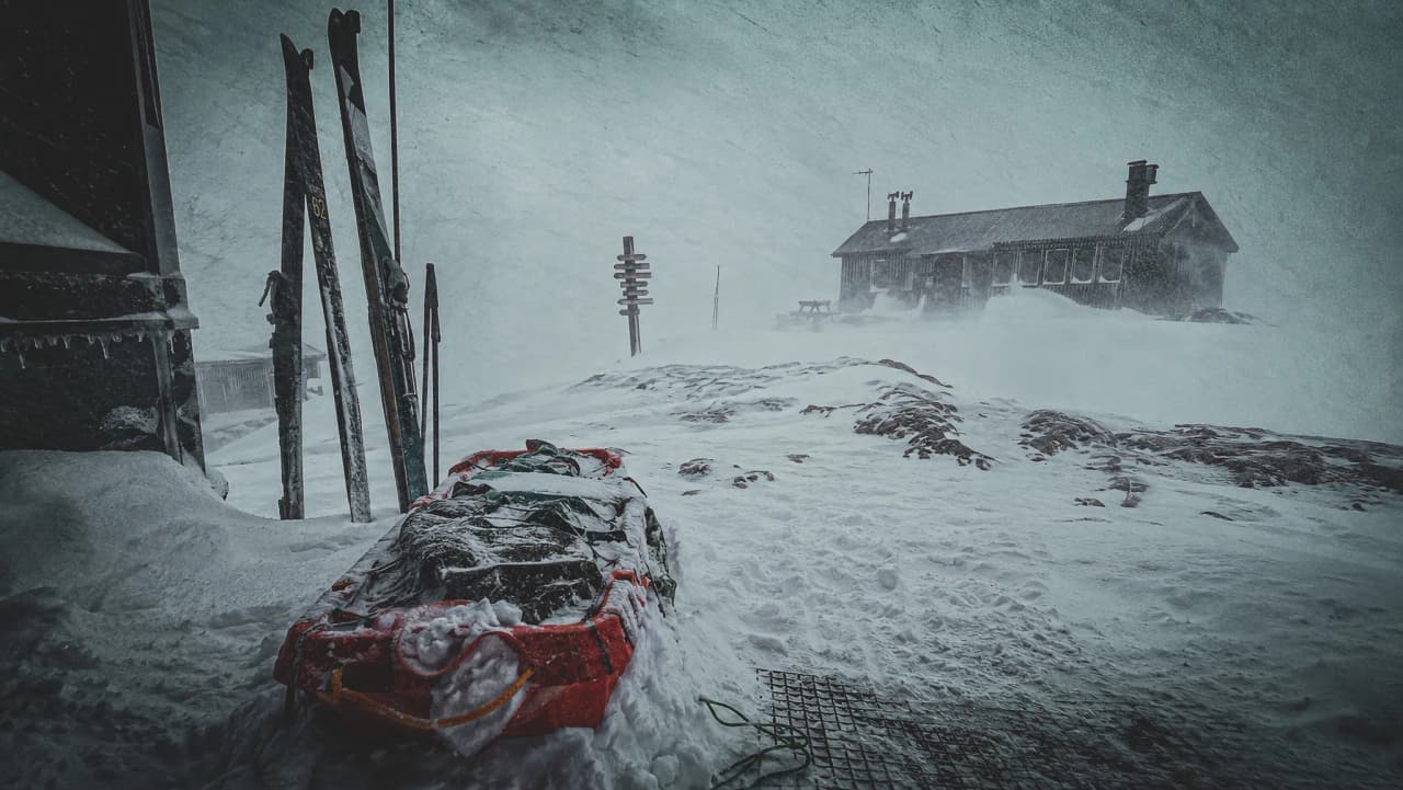 Een besneeuwd landschap langs de Kungsleden, met ski’s, een pulka en een Berghut tijdens een winterstorm.