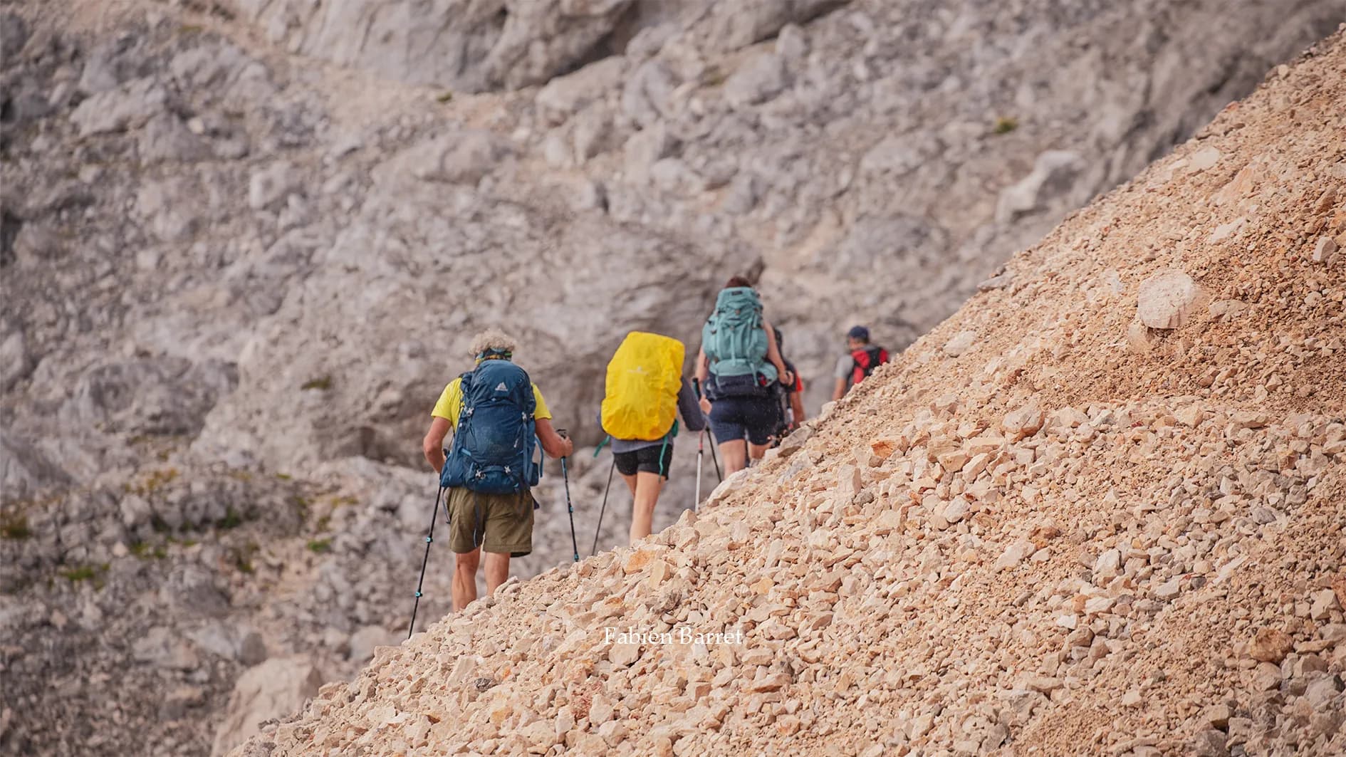 A majestic mountain landscape with steep, rocky peaks under a cloudy sky. In the foreground, a hiker equipped with poles and a rucksack makes his way across a terrain of rock and stone.