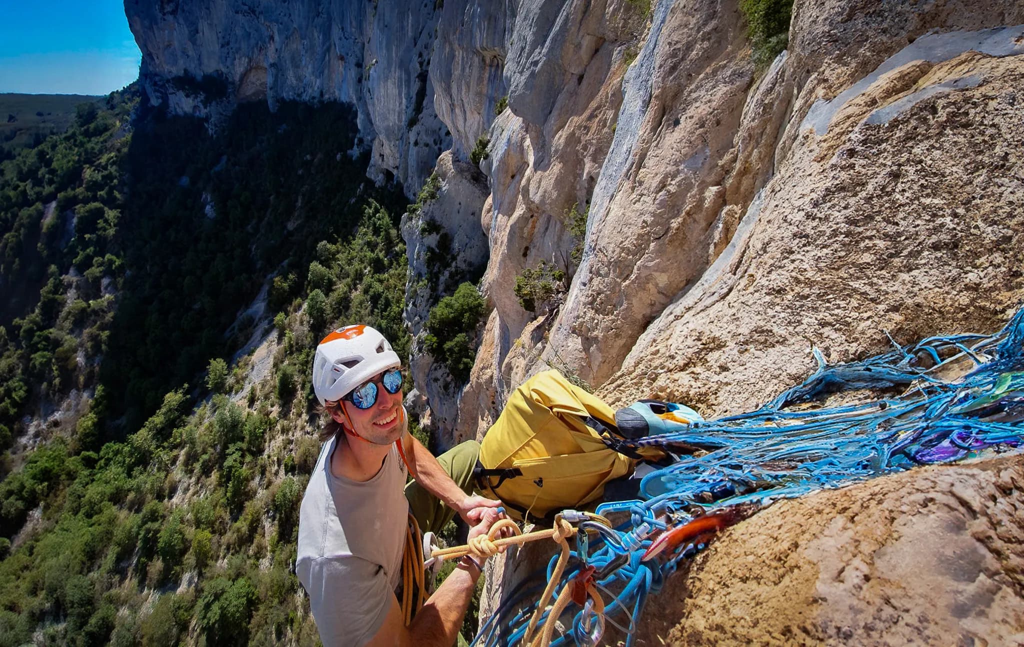 A climber is scaling a steep rock face, with a green landscape in the background. He is wearing a safety helmet and sunglasses, with a smile on his face. From