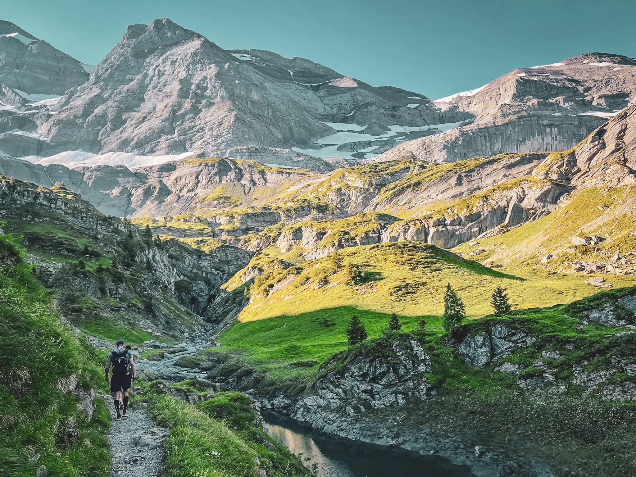 A hiking trail winding through spectacular mountain scenery, with craggy peaks and rock faces. Lush vegetation covers the slopes, while a tranquil stream flows past.