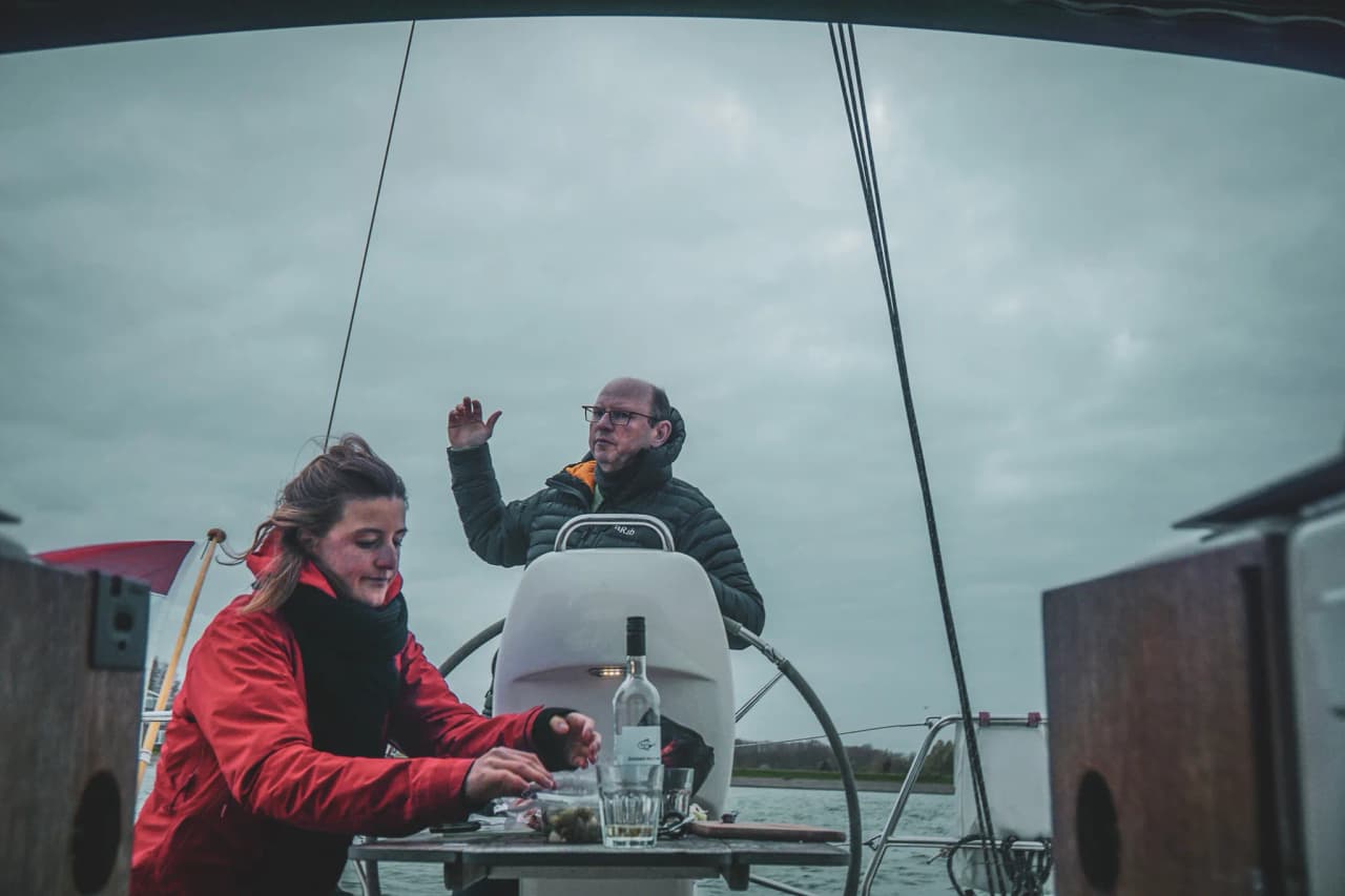 Een glimlachende vrouw maakt een snack klaar op een zeilboot, terwijl een man de weg wijst onder een bewolkte hemel.