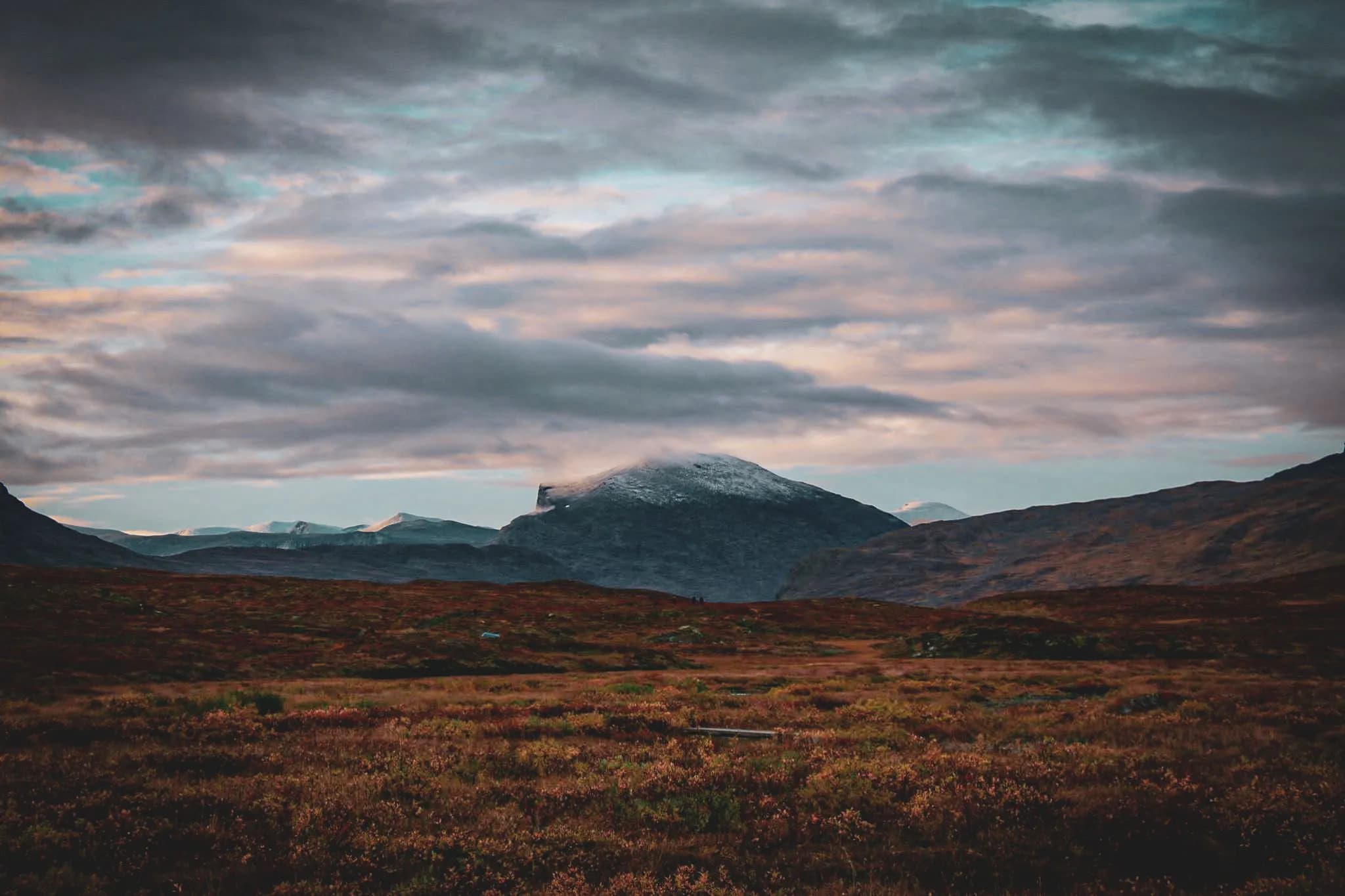 A mountainous landscape with majestic mountains in the background, some covered in snow. The sky is partly cloudy, with shades of blue and pastel hues. In the foreground, a vast plain