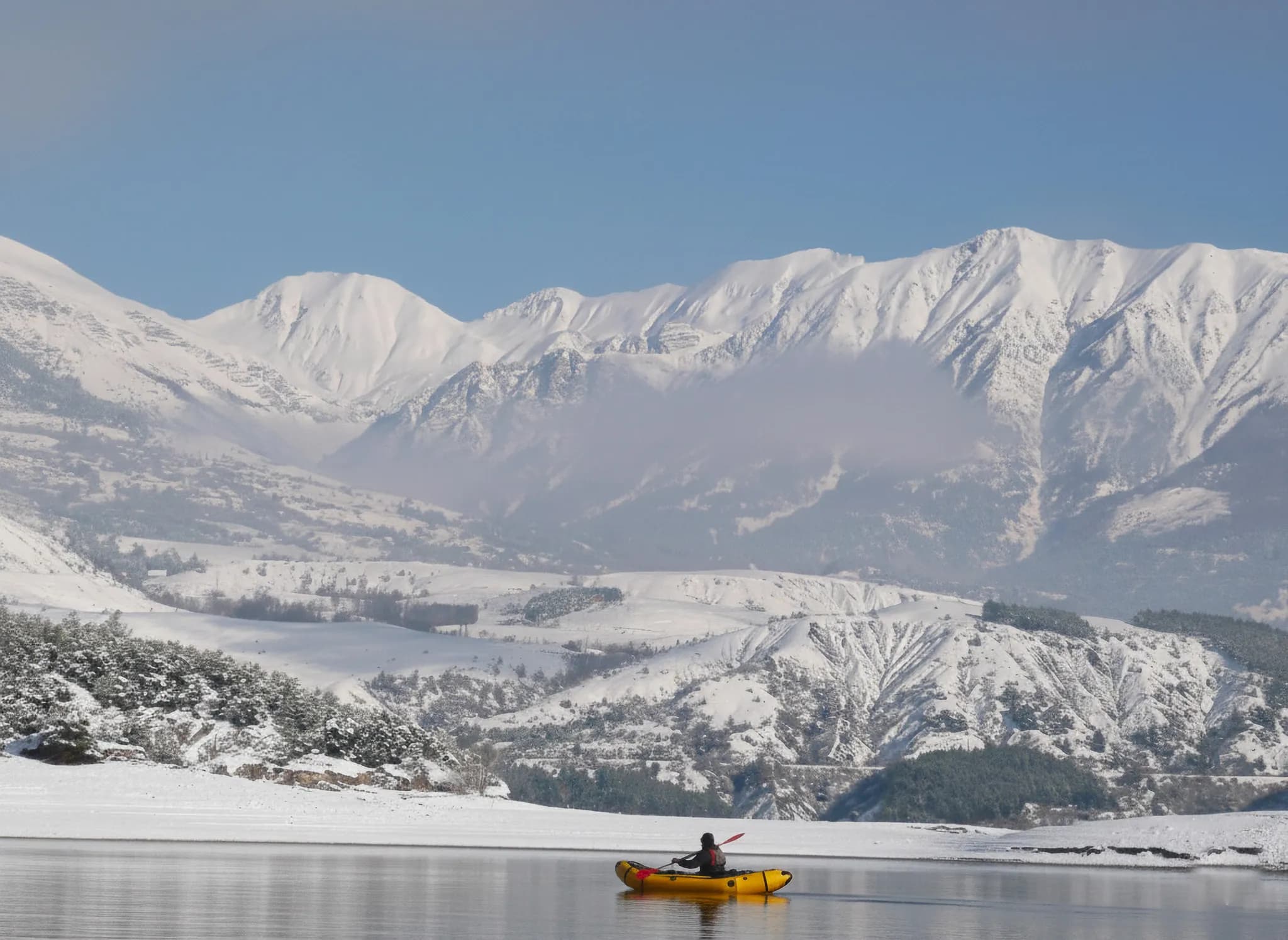 Un kayakiste navigue sur le lac de Serre-Ponçon, entouré de montagnes enneigées et d'un ciel bleu.