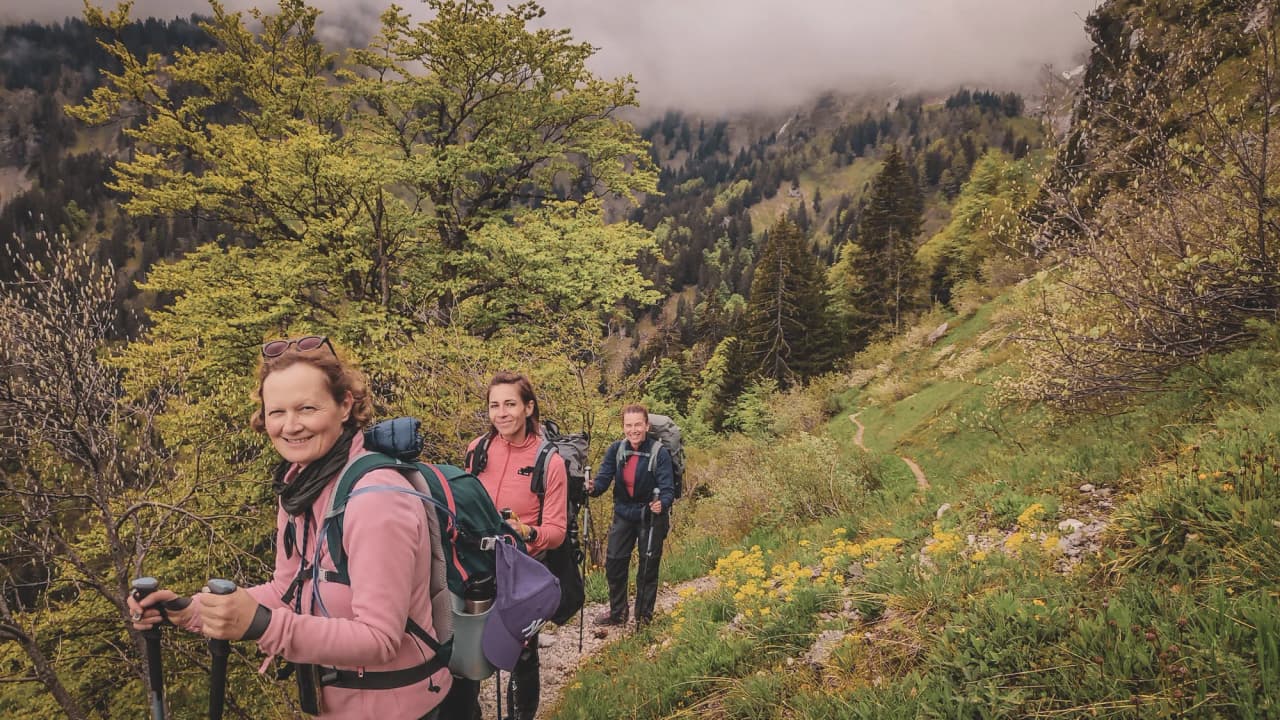 Three smiling hikers on a green trail, with majestic peaks in the background.