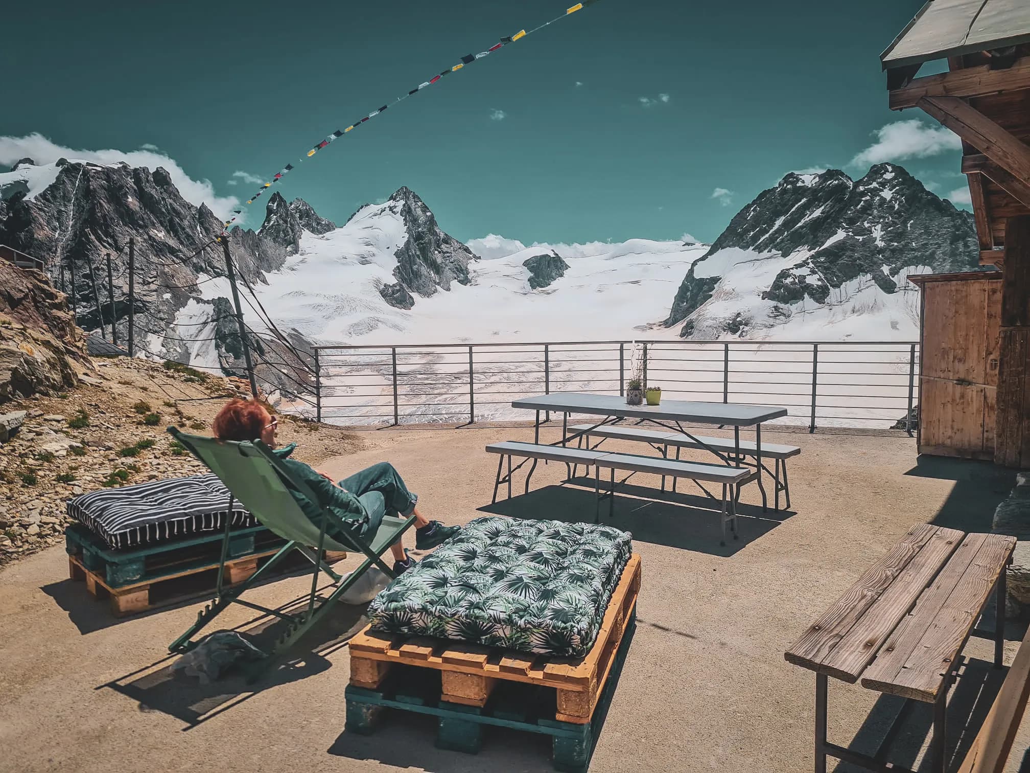 A high-altitude terrace with a breathtaking view of majestic mountain landscapes, marked by snow-capped peaks. A deckchair is positioned in front of a picnic table, surrounded by colourful mattresses.