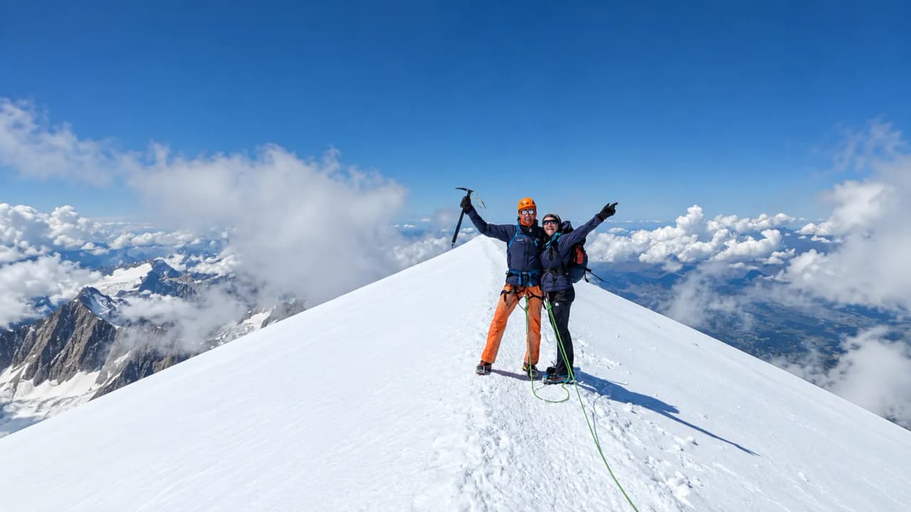 Deux alpinistes au sommet du Mont Blanc, rayonnants, entourés de montagnes majestueuses et de ciel bleu.