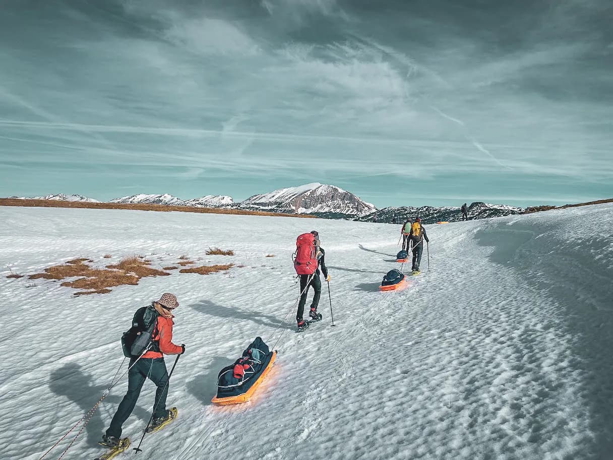 A group of hikers venture across a snowy landscape, pulling sledges behind them. They are dressed in colourful outdoor clothing, with well-filled rucksacks. In the background,