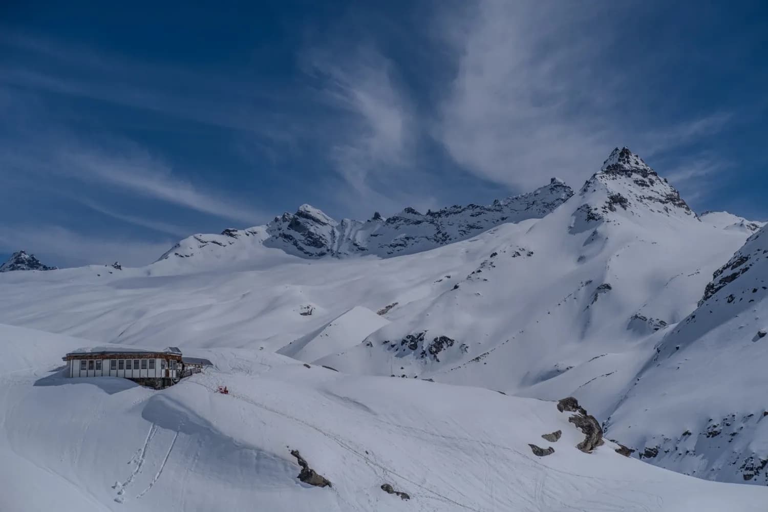A snow-covered Alpine landscape, dominated by majestic peaks under a blue sky with scattered clouds. In the foreground, a modern chalet contrasts harmoniously with the whiteness of the surrounding snow. Traces of