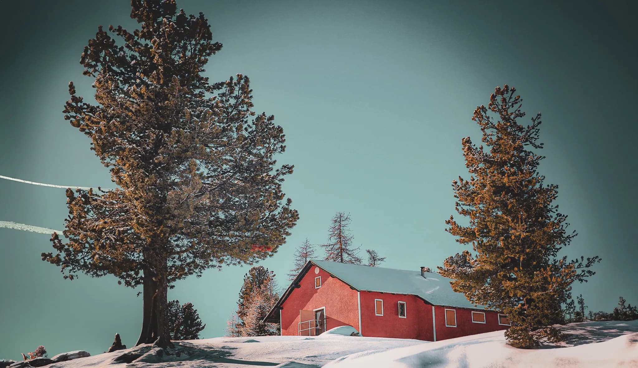 A red chalet is nestled in a snowy landscape, surrounded by tall green trees. The sky is a clear blue, with white streaks of aircraft crossing the horizon. The snow is rec