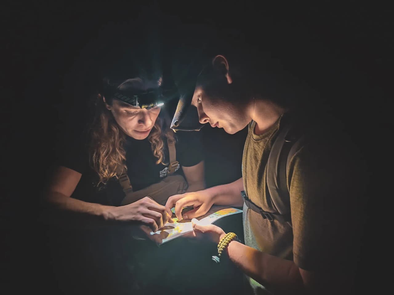 Deux personnes éclairées par des lampes frontales étudient une carte dans l'obscurité, prêtes à explorer.