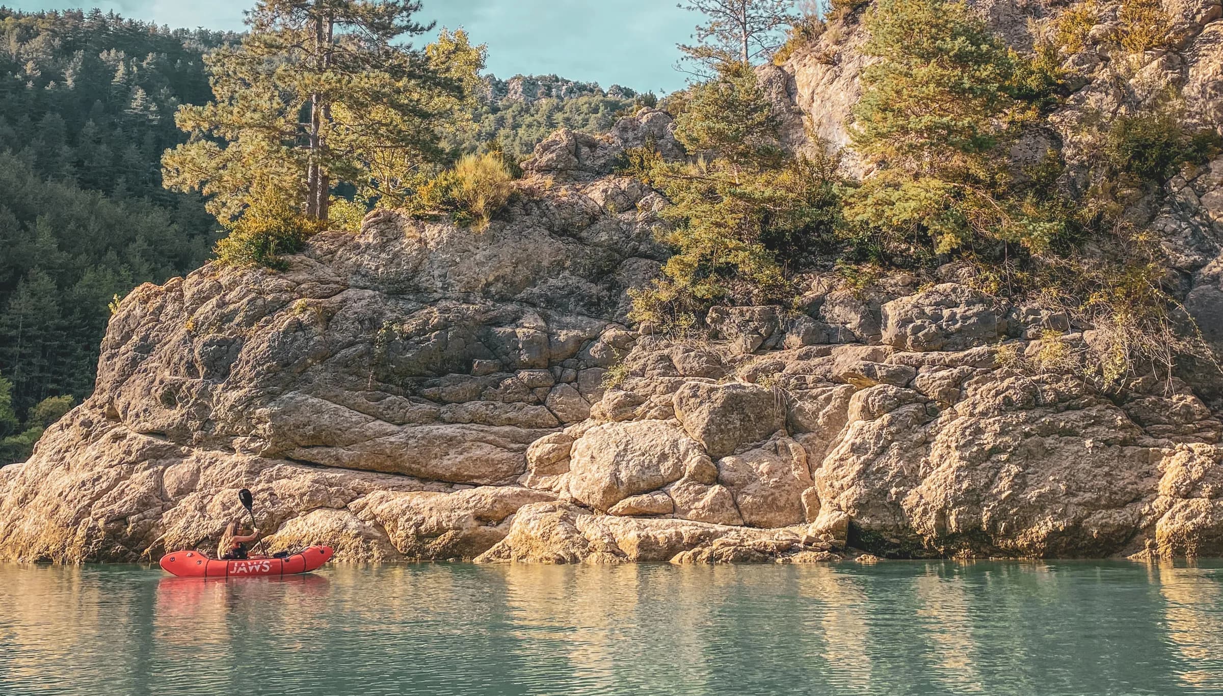 A red kayak floating peacefully on calm water, close to a large rock formation on the edge of a lake. Lush green trees stand both on the rock and behind it, creating a natural and tranquil atmosphere.