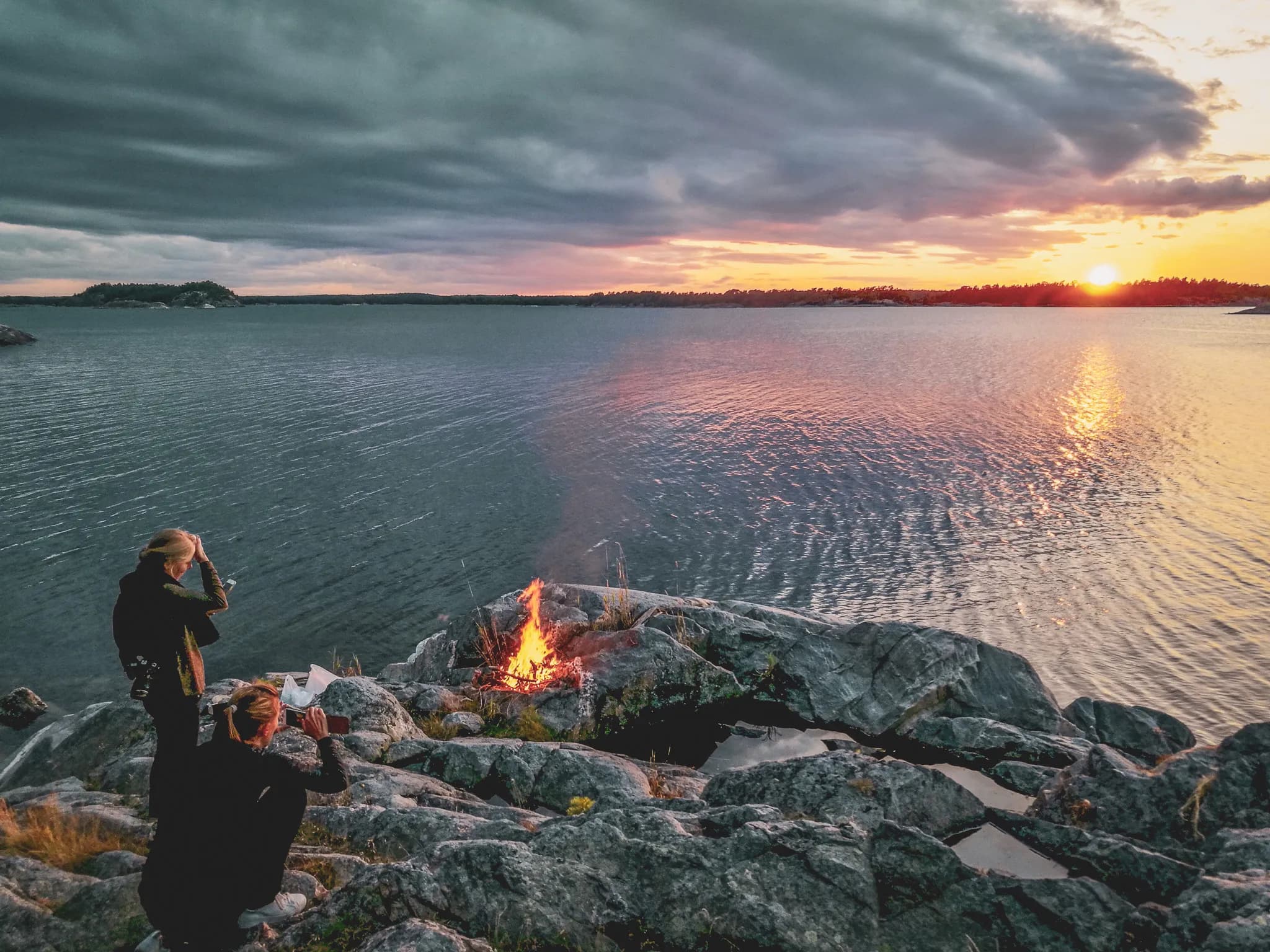 A spectacular sunset over a lake, with dark clouds and reflections of light on the water. Two people are standing by a fire on some rocks, one of them taking a photo. The setting is