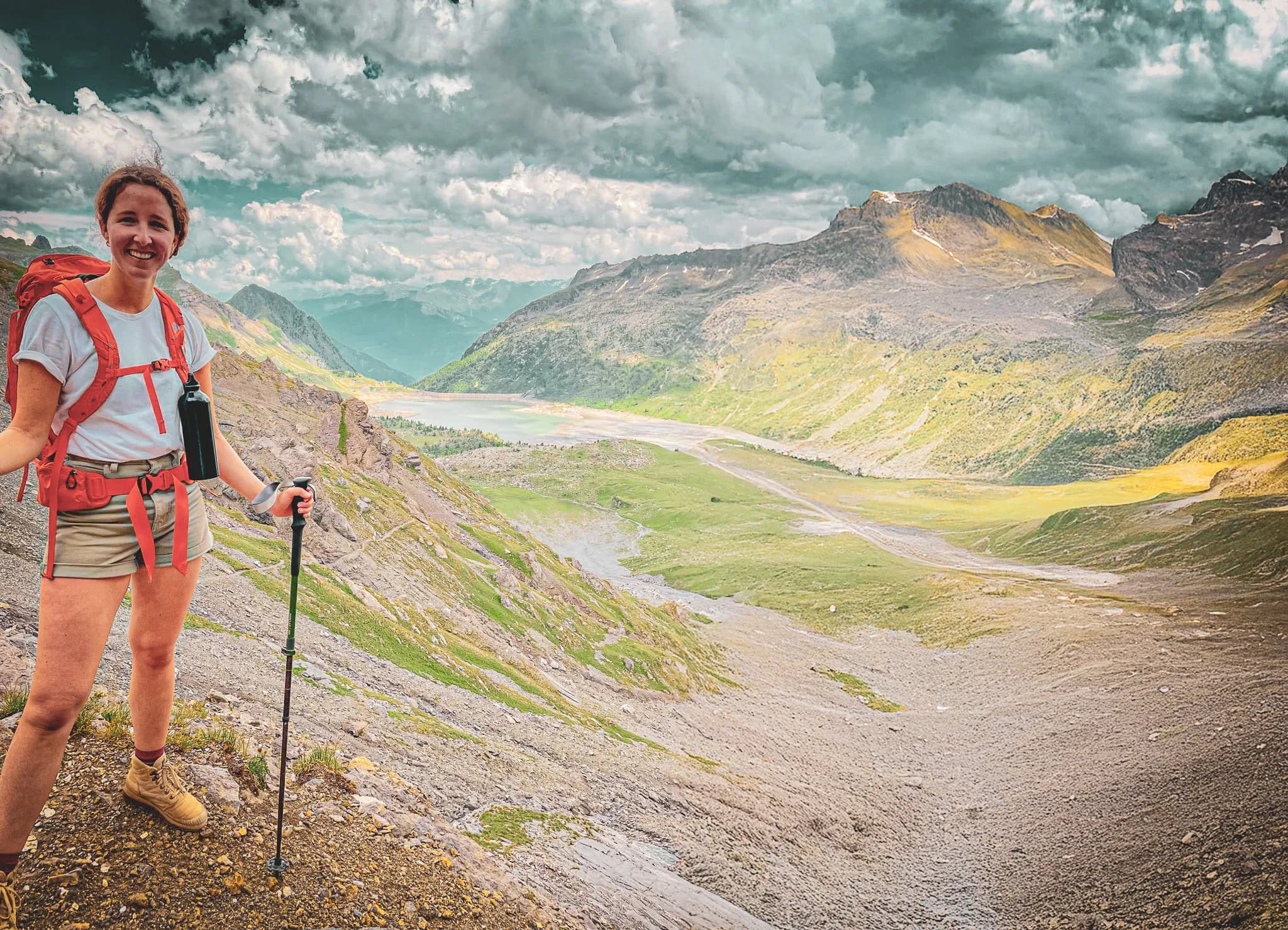 A hiker strikes a smiling pose on a mountain path, with a landscape of green valleys and mountains in the background. The sky is cloudy, with shades of grey and blue.