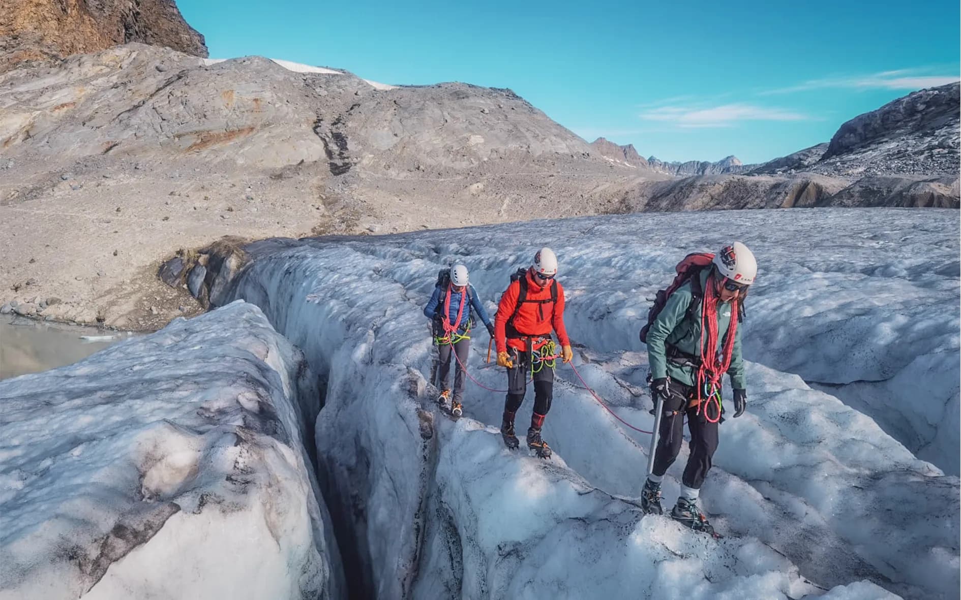 A vast, bright glacier, with visible cracks, stretches out beneath a clear blue sky. Three climbers, equipped with helmets and ropes, advance cautiously across the icy surface. The first,