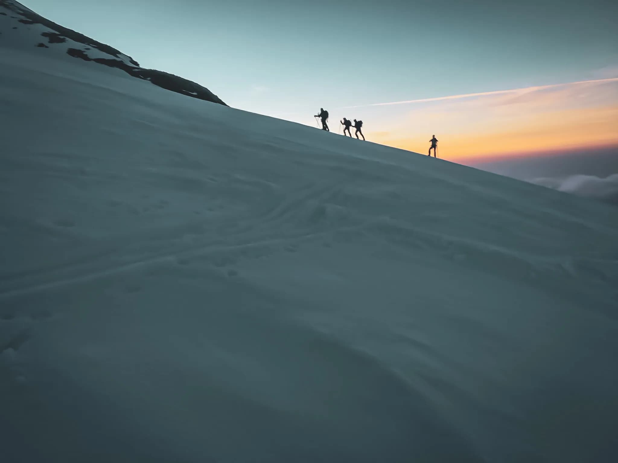 Een besneeuwd berglandschap, waar verschillende silhouetten van wandelaars oprijzen tegen een helling in de schemering. De lucht is getint met pasteltinten, veranderend van blauw naar geeloranje, terwijl een deel van de lucht bedekt is met sneeuw.