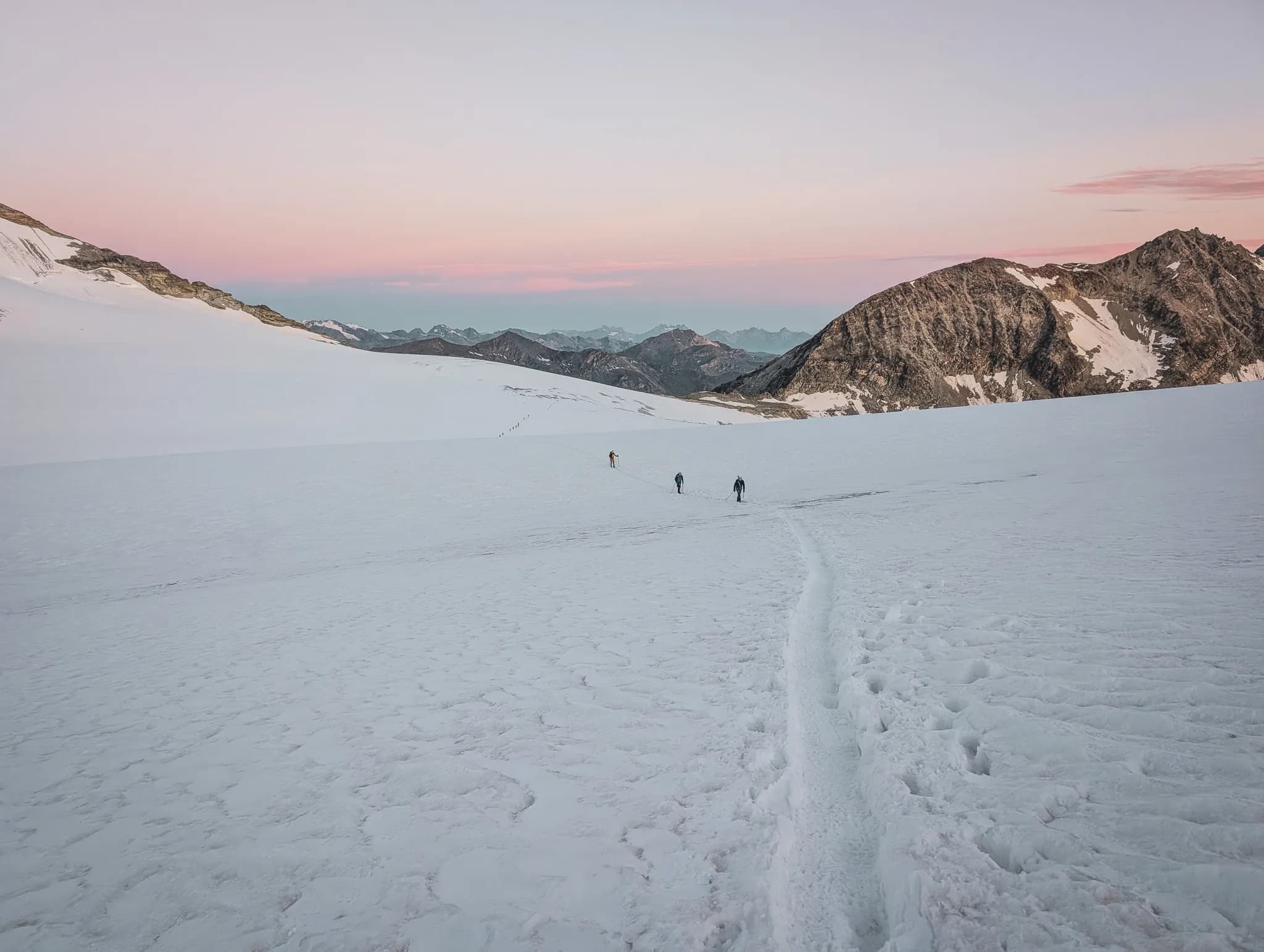 A spectacular mountain landscape unfolds beneath a sky tinged with pink and purple hues at dusk. Snow-capped peaks rise majestically, some with rocky slopes.