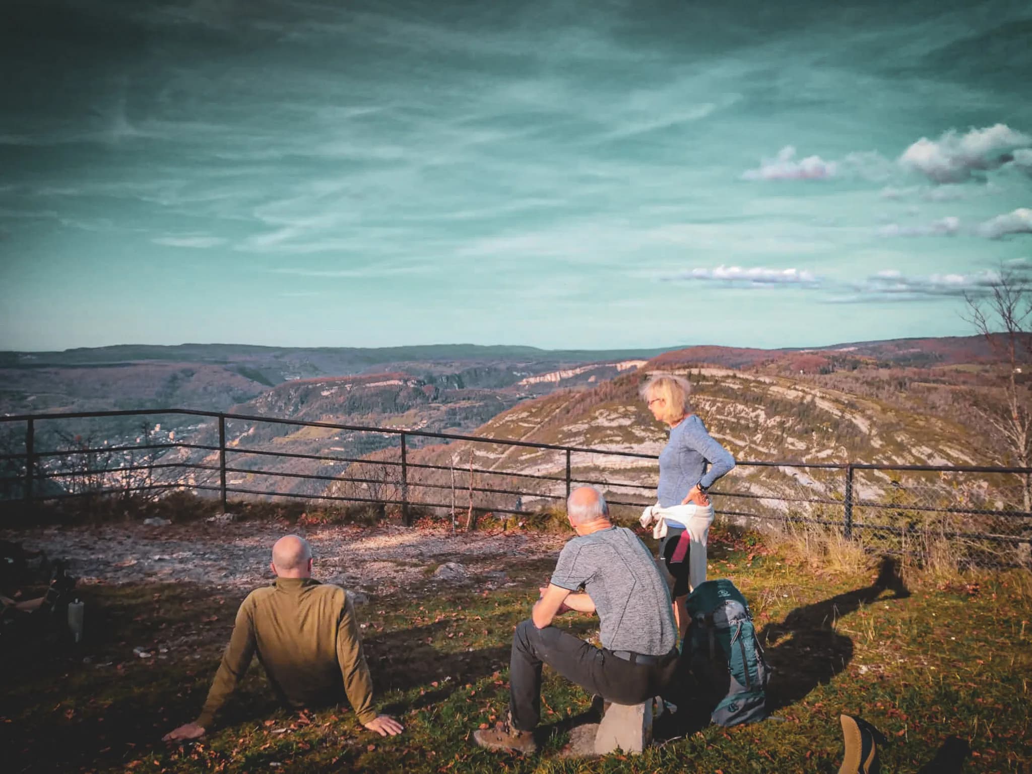 Three people are enjoying a panoramic view of a valley. Two men are sitting on the ground, one facing the stage, the other looking in another direction. A woman is standing,