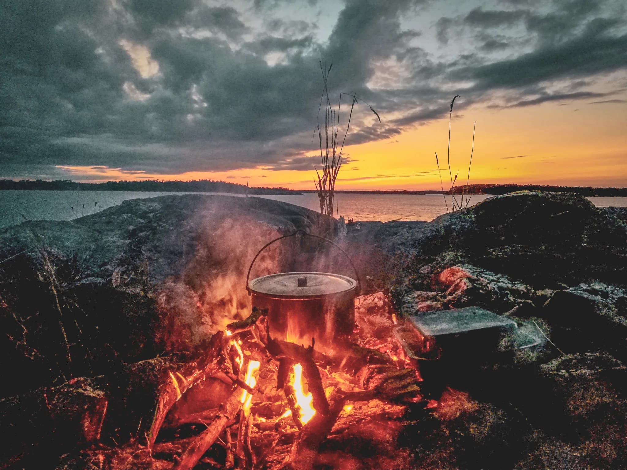 A campfire crackles on a large stone on the edge of a lake, with a metal pot sitting on the fire. The light from the flames illuminates the scene, while a light smoke rises into the air. Visit