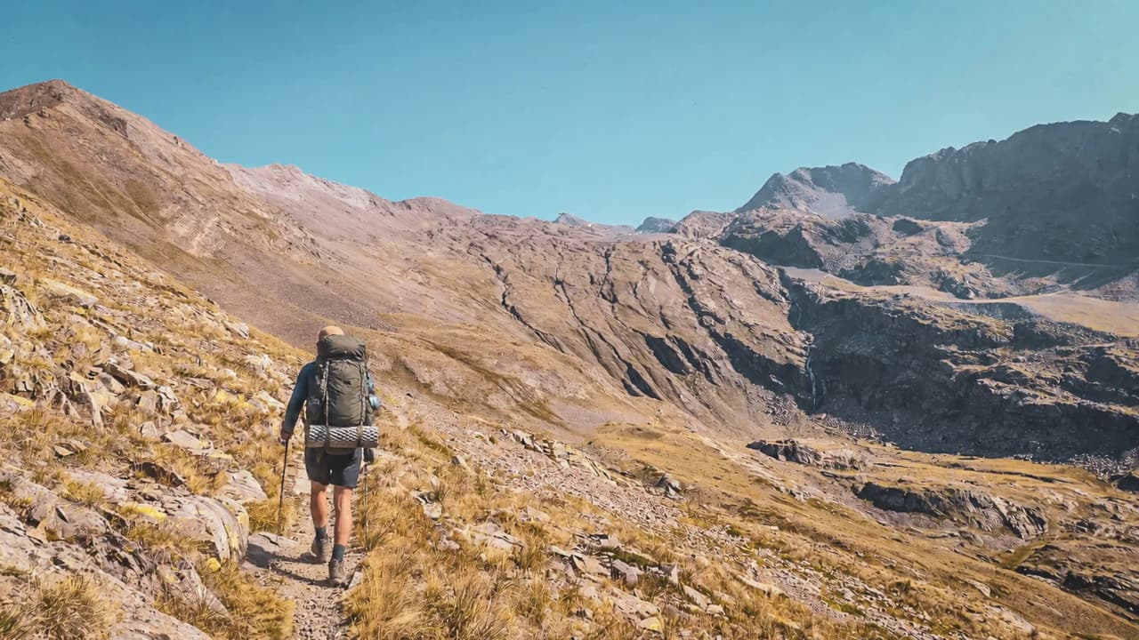 Randonneur traversant un paysage alpin aride, avec de majestueux sommets en toile de fond.