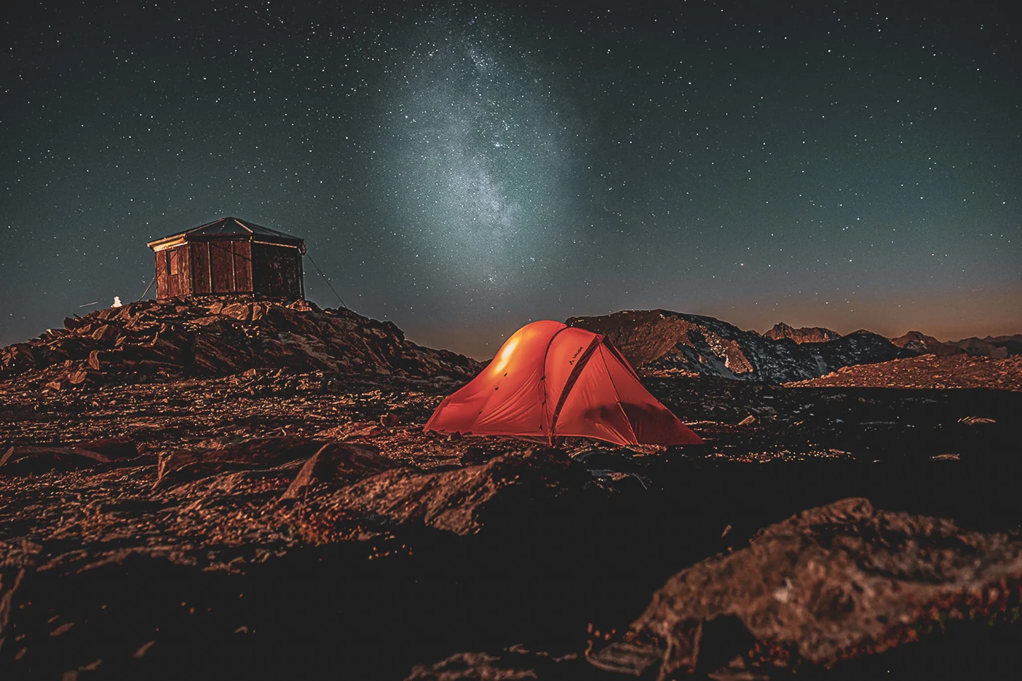 A mountain nightscape with a shiny red tent set in rocky terrain. Nearby, a small wooden building with a sloping roof stands on a hill. The sky is full of stars, and a