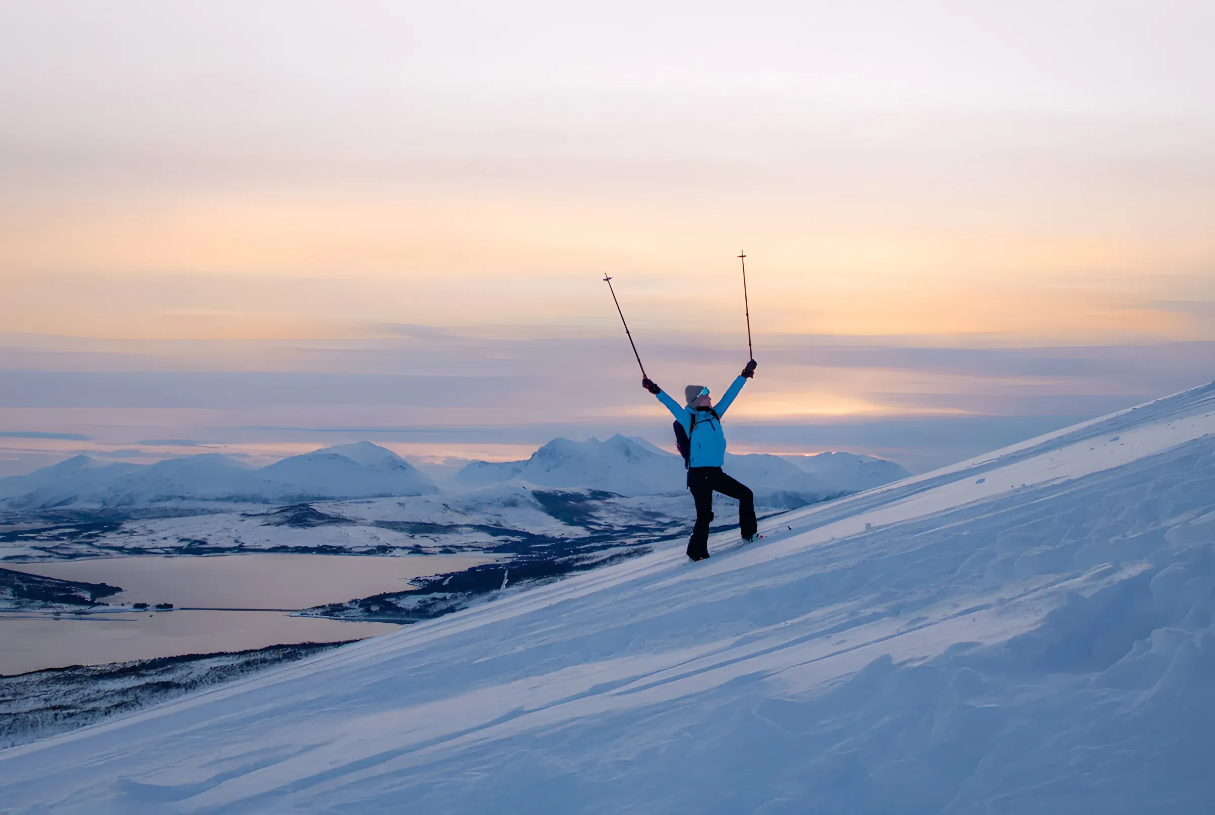 A person on snow-covered slopes lifting their poles under a colourful sky at dusk on the island of Senja.