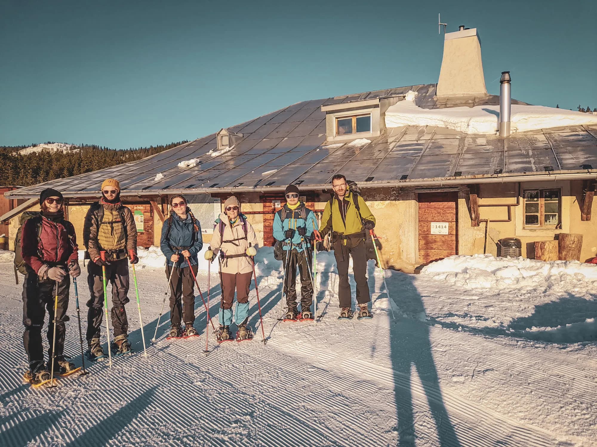 Groupe d'aventuriers en raquettes devant un refuge enneigé dans le Jura suisse, sous un ciel clair.