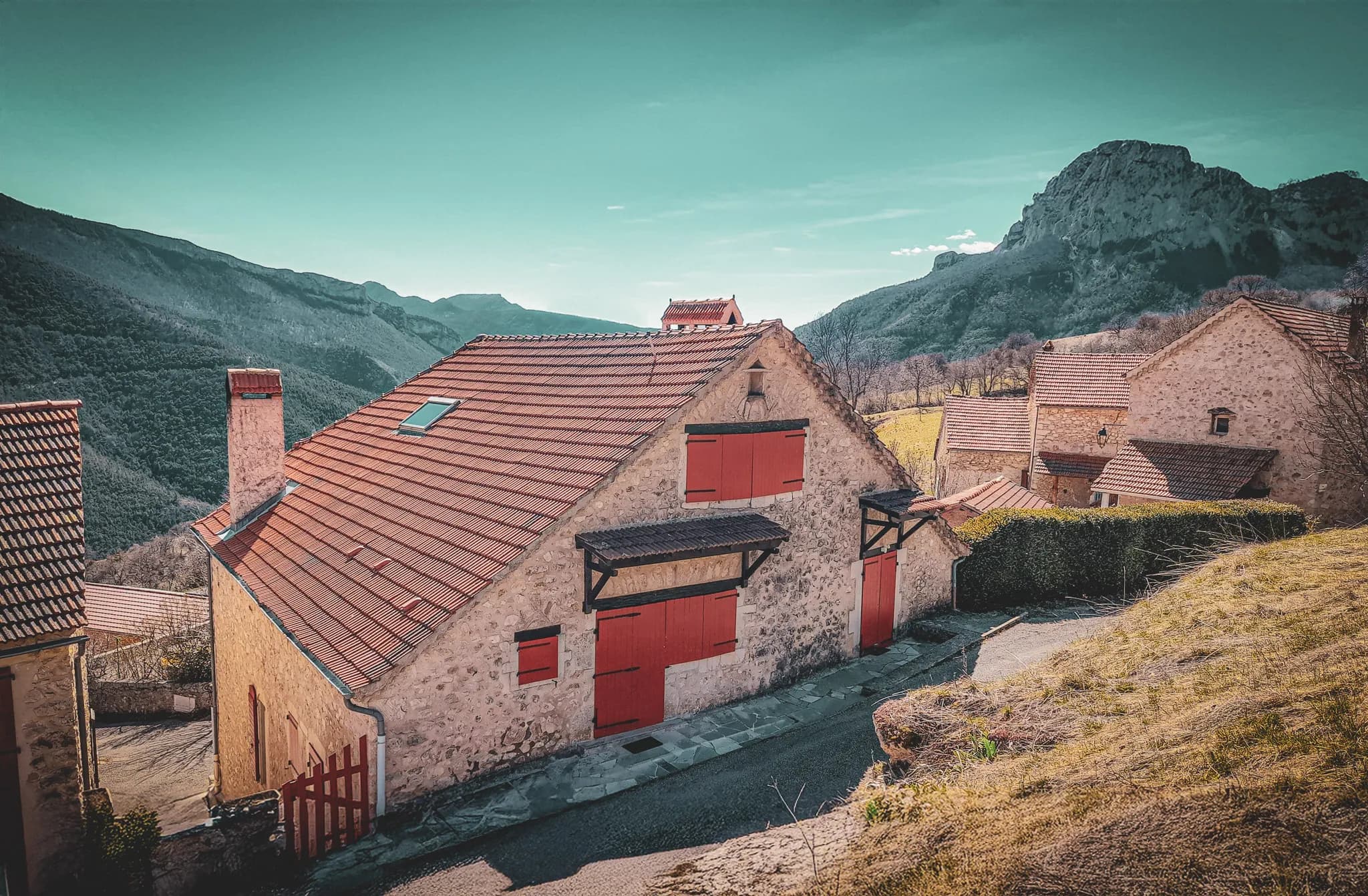 A charming stone cottage with a red-tiled roof, set in a mountain landscape. The front of the building features red shutters and wooden architectural details. In the background, lush green mountains