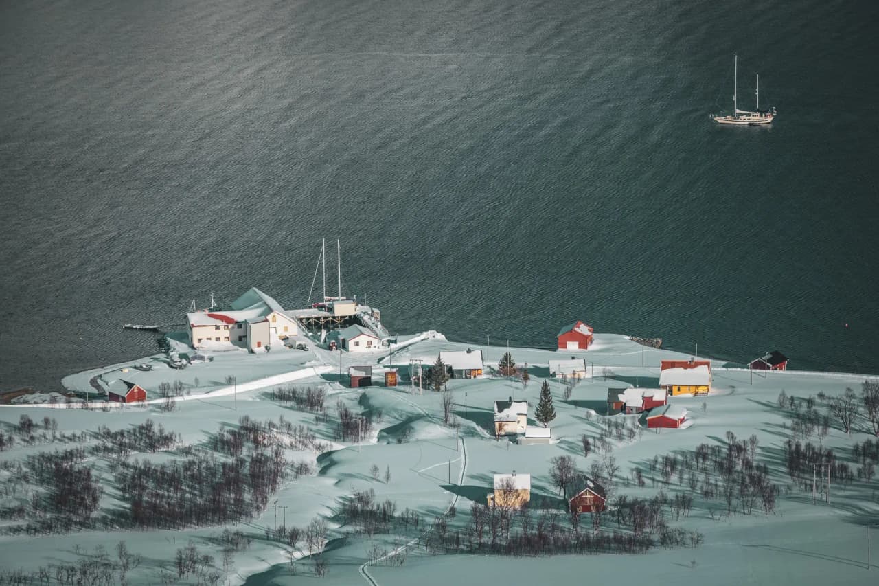 A picturesque snow-covered village overlooking a fjord, with colourful houses and a sailboat.
