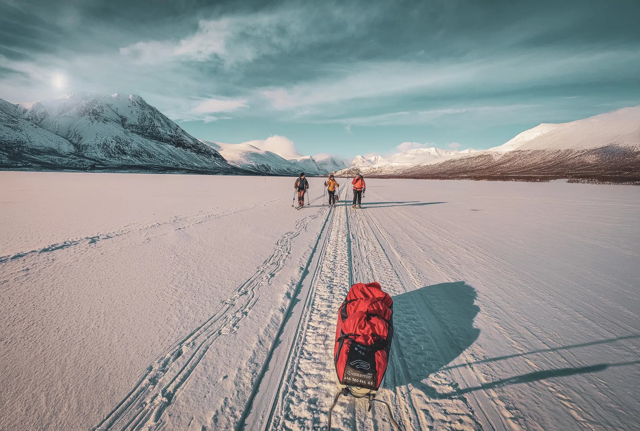 A snow-covered mountain landscape, with several hikers walking on a frozen surface. They are pulling a red pulka behind them. Sunlight shines on the mountains in the distance.