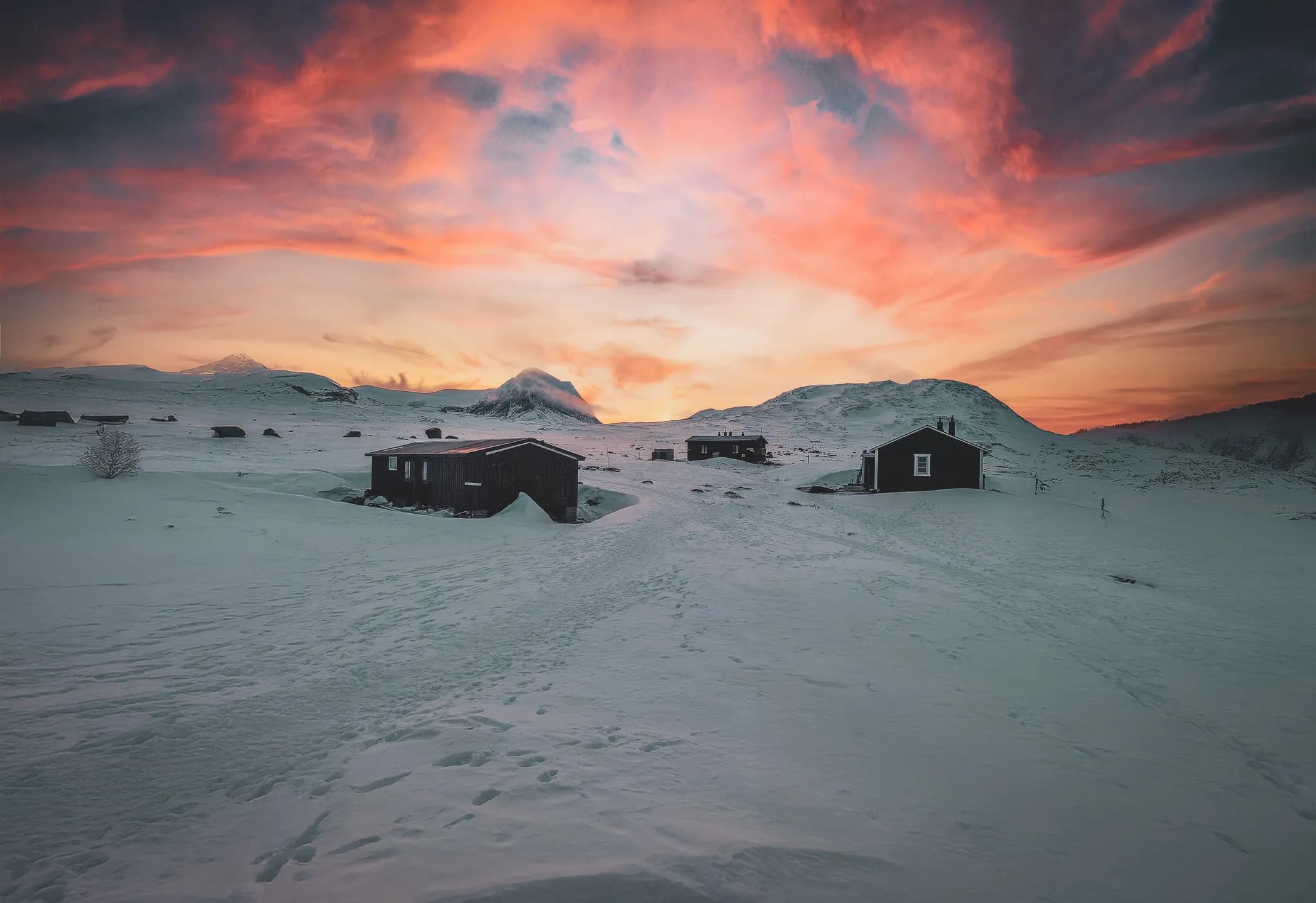 The winter landscape of Kungsleden with several wooden chalets surrounded by immaculate snow. The sky is tinged with shades of orange and pink, creating a striking contrast with the whiteness of the ground.