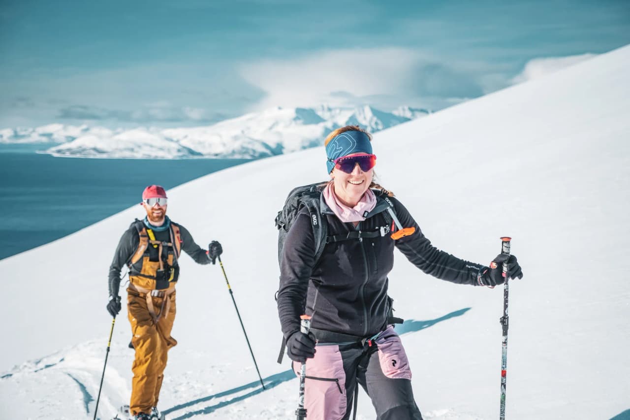 Two people hiking in a snowy alpine landscape, with majestic mountains in the background. One of them, smiling, is wearing a black jacket and pink ski trousers, as well as a headband and goggles.