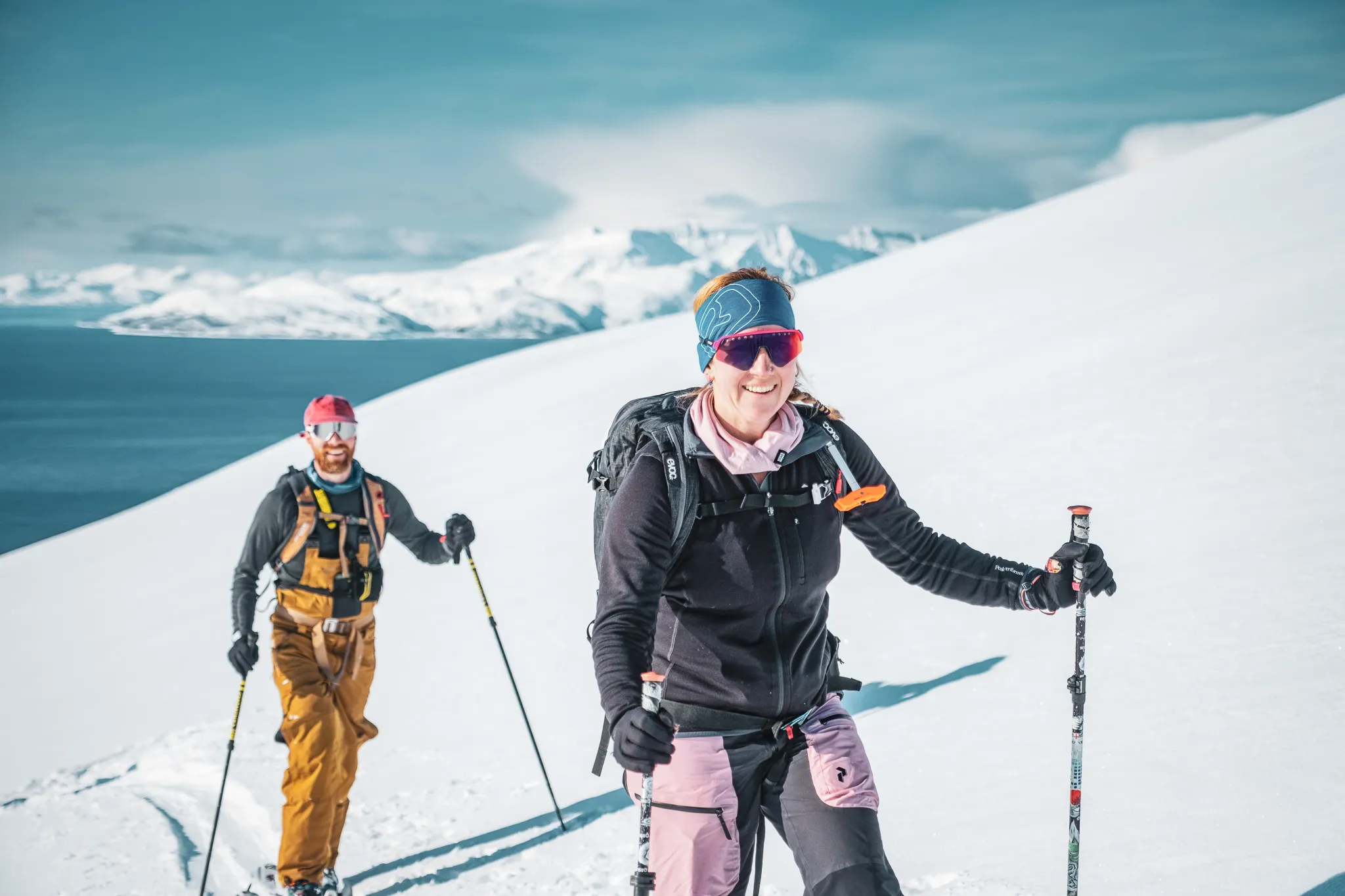 Deux personnes en randonnée dans un paysage alpin enneigé, avec des montagnes majestueuses en arrière-plan. L'une des personnes, souriante, porte une veste noire et un pantalon de ski rose, ainsi qu'un bandeau et des lunettes
