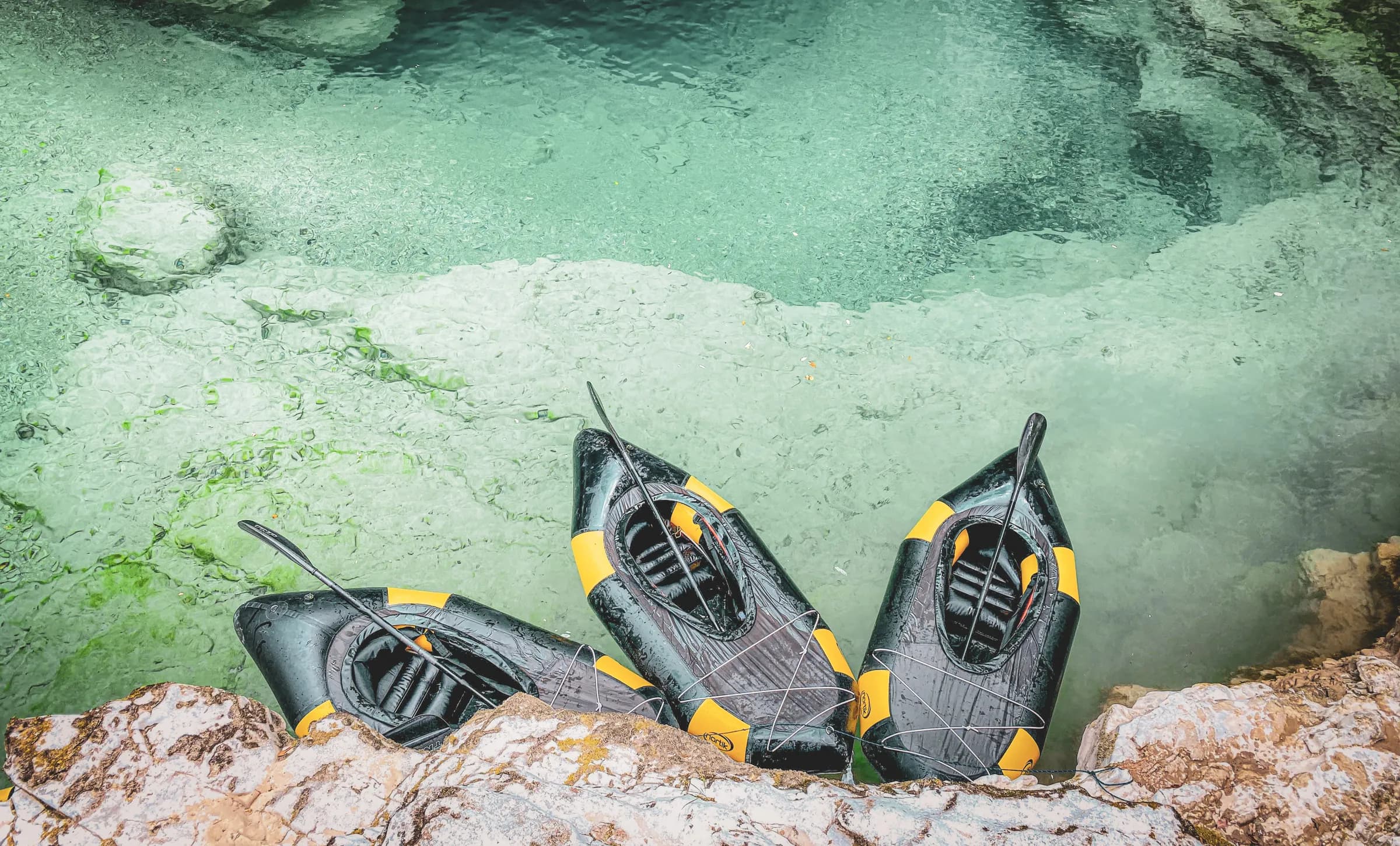 A natural setting surrounded by rock formations, where several black kayaks with yellow edges are moored on the edge of clear, calm water. The emerald-green water reveals rocky bottoms and