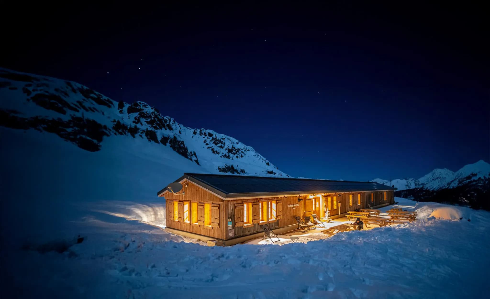 Un chalet en bois se dresse dans un paysage enneigé, illuminé par la lumière chaude de ses fenêtres, créant une ambiance accueillante au milieu de la nuit. La structure est entourée de neige immaculée, tandis que des