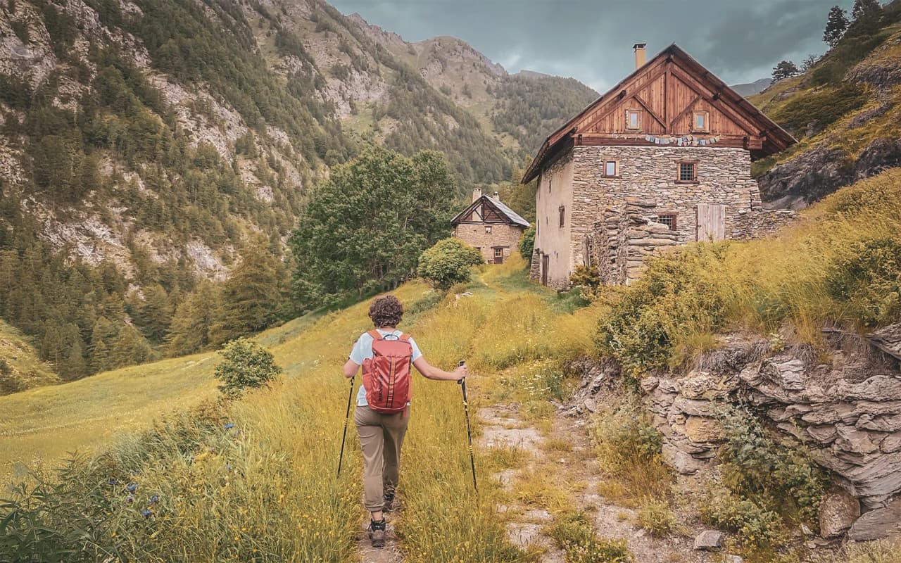 Hiker on a green path, facing typical Queyras chalets in the surrounding mountains.