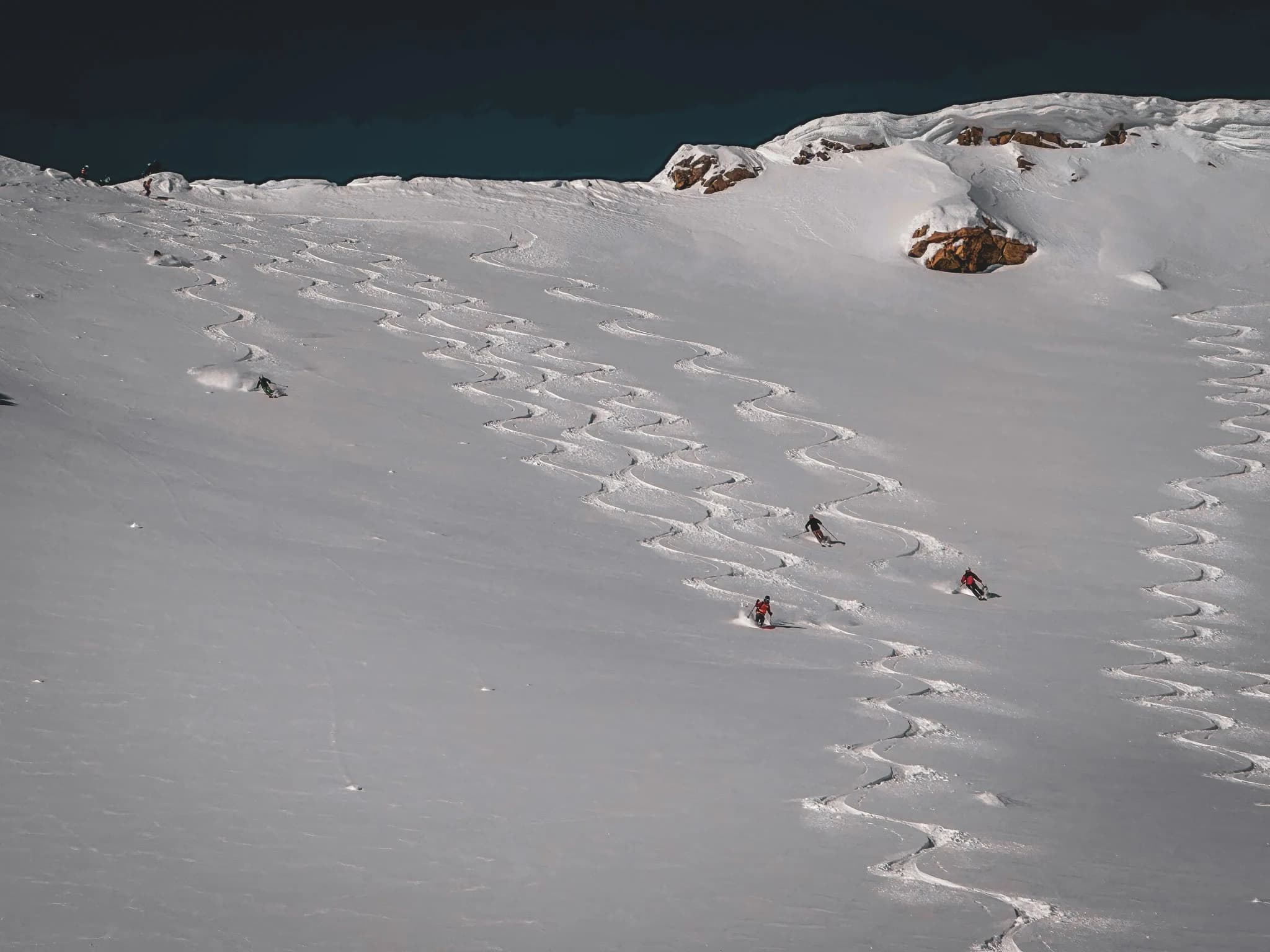 Une vaste étendue de neige immaculée, marquée par des traces de ski sinueuses. Plusieurs skieurs descendent la pente, laissant derrière eux des sillons qui serpentent sur la surface enneigée. Le contraste entre le blanc