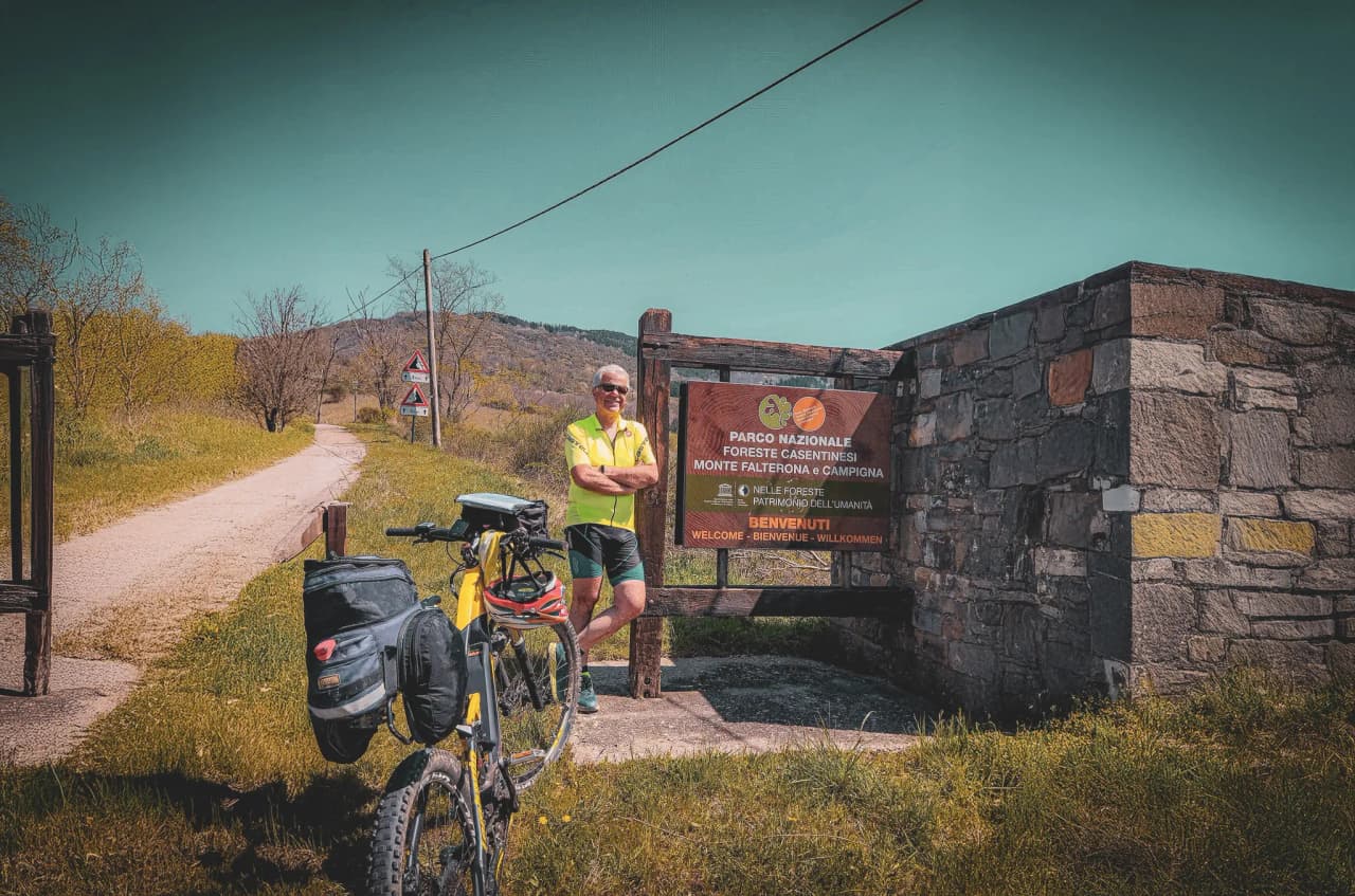 A man stands beside his bike, smiling, in front of a stone entrance to a national park. Wearing a bright yellow jersey and cycling shorts, he looks ready to explore nature.