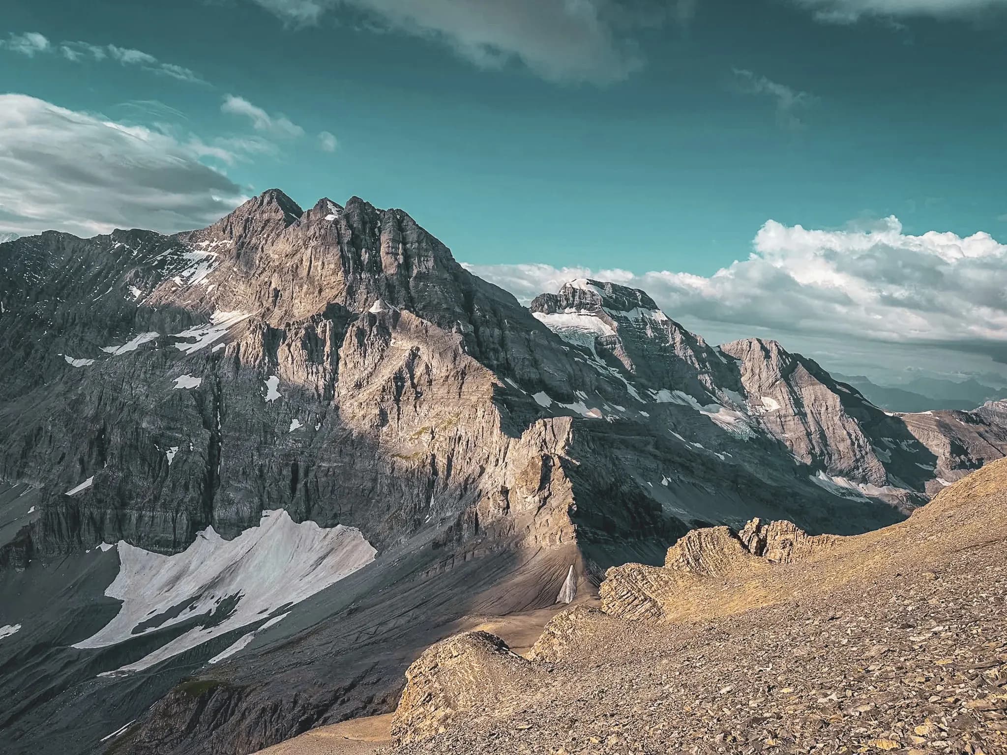 A spectacular mountain landscape, characterised by steep peaks and well-defined rocky ridges, with shades of grey and brown, interspersed with traces of snow. The sky is partly cloudy