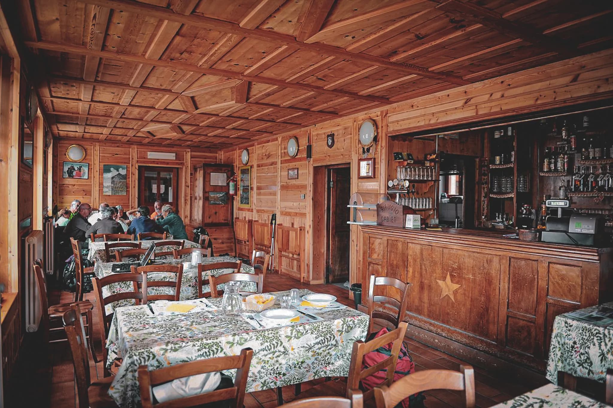 Een warm en gastvrij interieur kenmerkt een houten restaurant met gelambriseerde muren en plafonds. Er zijn verschillende tafels gedekt met gebloemde tafelkleden, sommige al gedekt met lege borden.