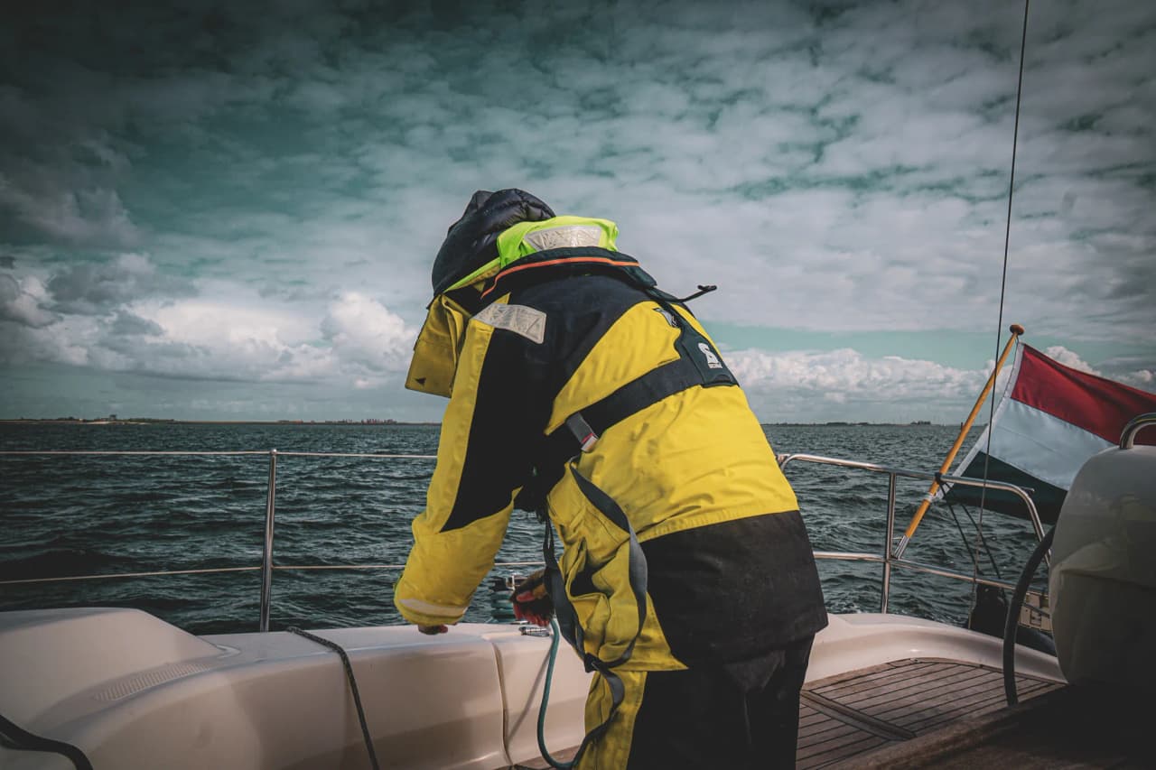Een matroos in gele overalls verstelt touwen op een zeilboot, met majestueuze wolken op de achtergrond.