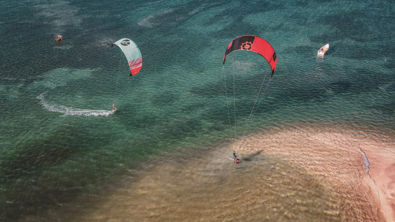 Kitesurf dans les eaux cristallines des Cyclades, avec des voiles colorées et des plages de sable.