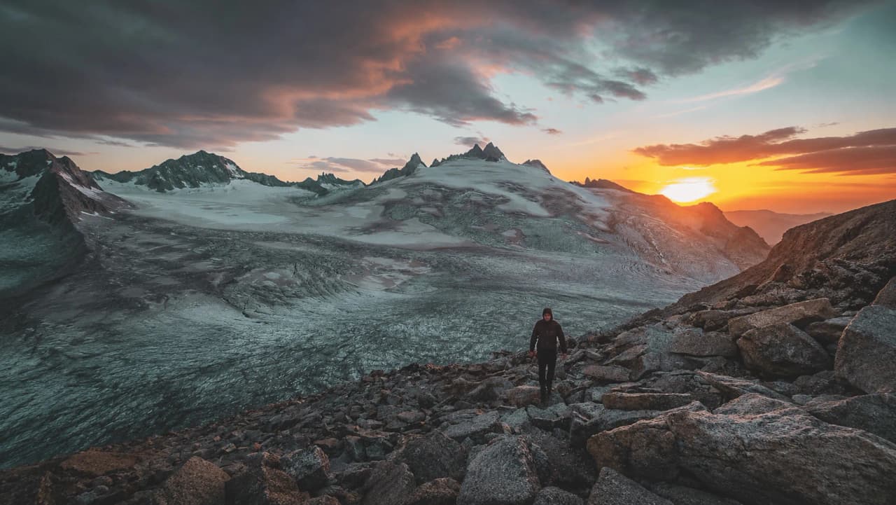 Randonneur sur des rochers face à un glacier au coucher de soleil, ambiance majestueuse.