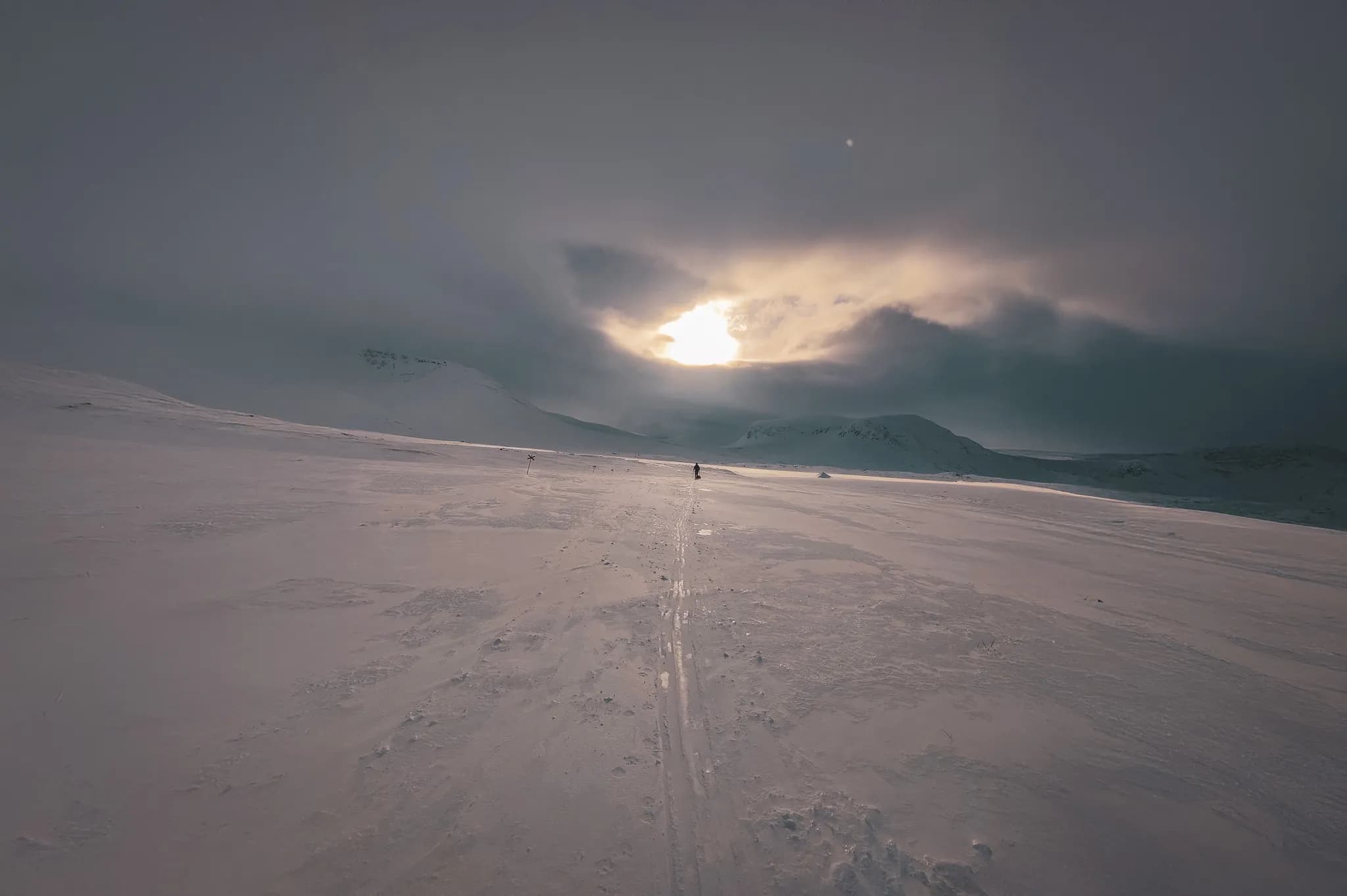 A vast Arctic winter landscape covered in brilliant snow stretches as far as the eye can see, with mountains looming on the horizon under a cloudy sky tinged with dark hues.