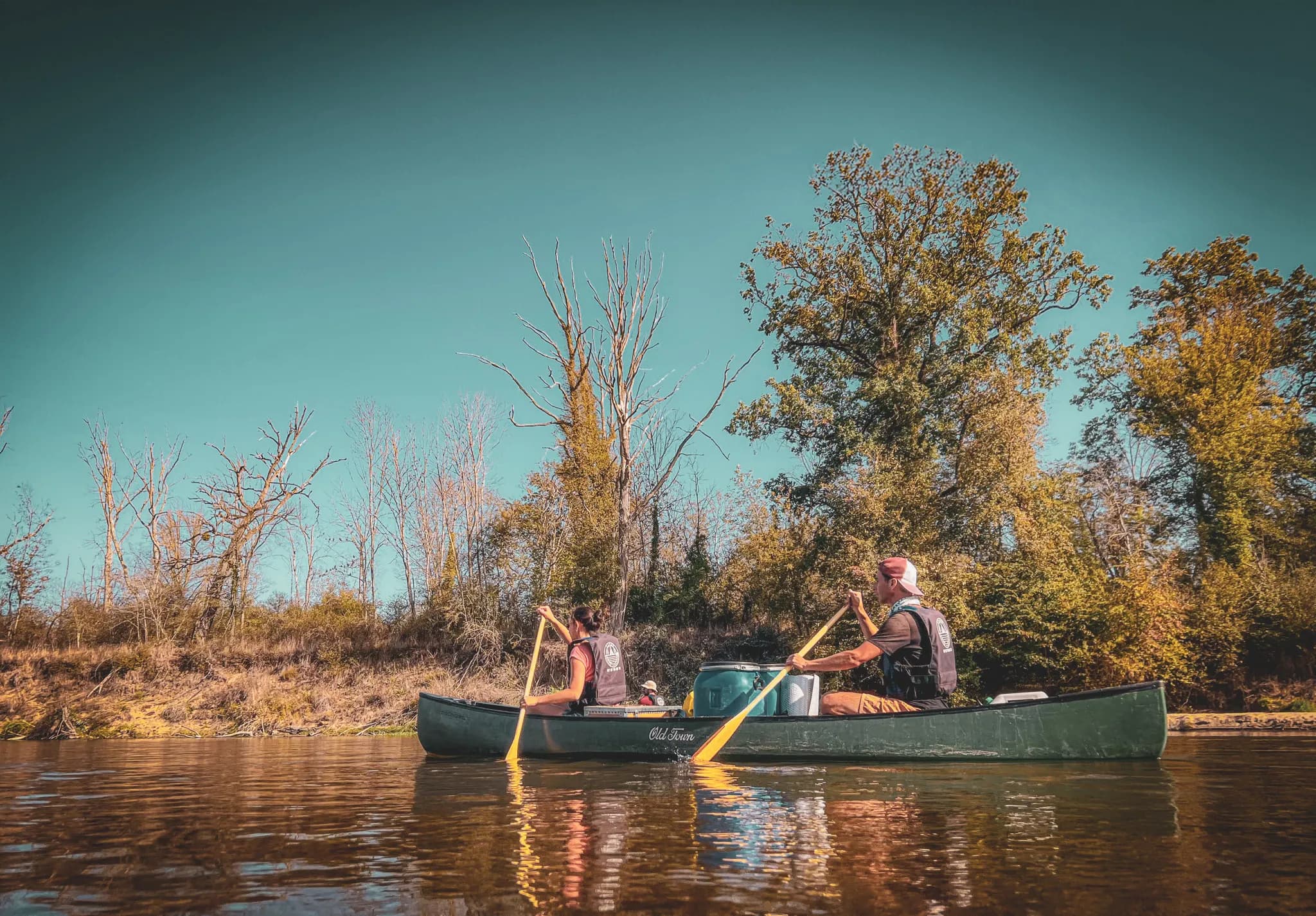 A canoe on a calm river, carried by two paddlers. The setting is natural, surrounded by trees with yellow and green foliage, a sign of autumn. The sky is clear