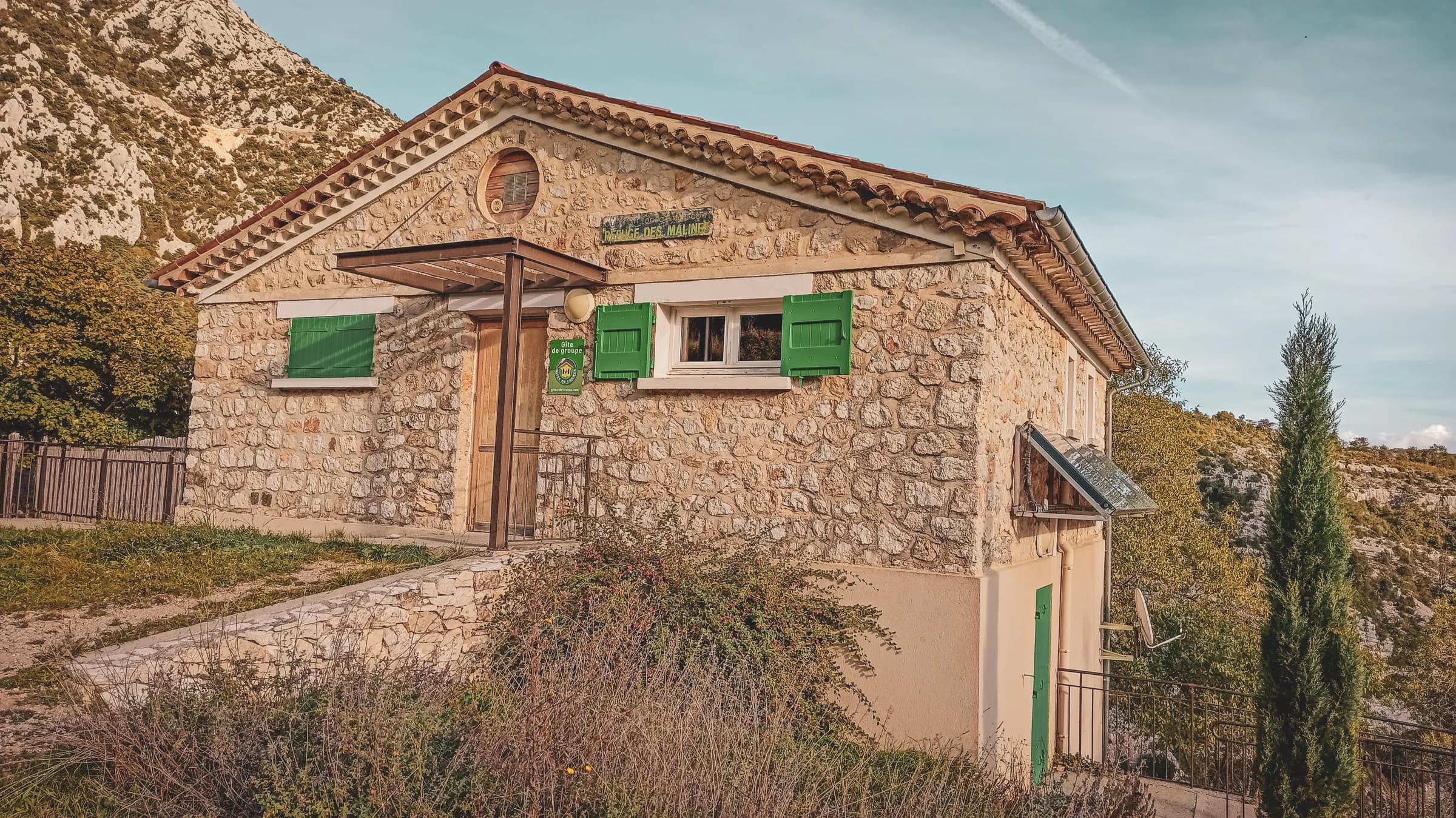A stone gîte in a natural setting, with green shutters and a tiled roof. The façade is stone, and the front door is wooden. Next to the gîte, plants and shrubs have been planted.